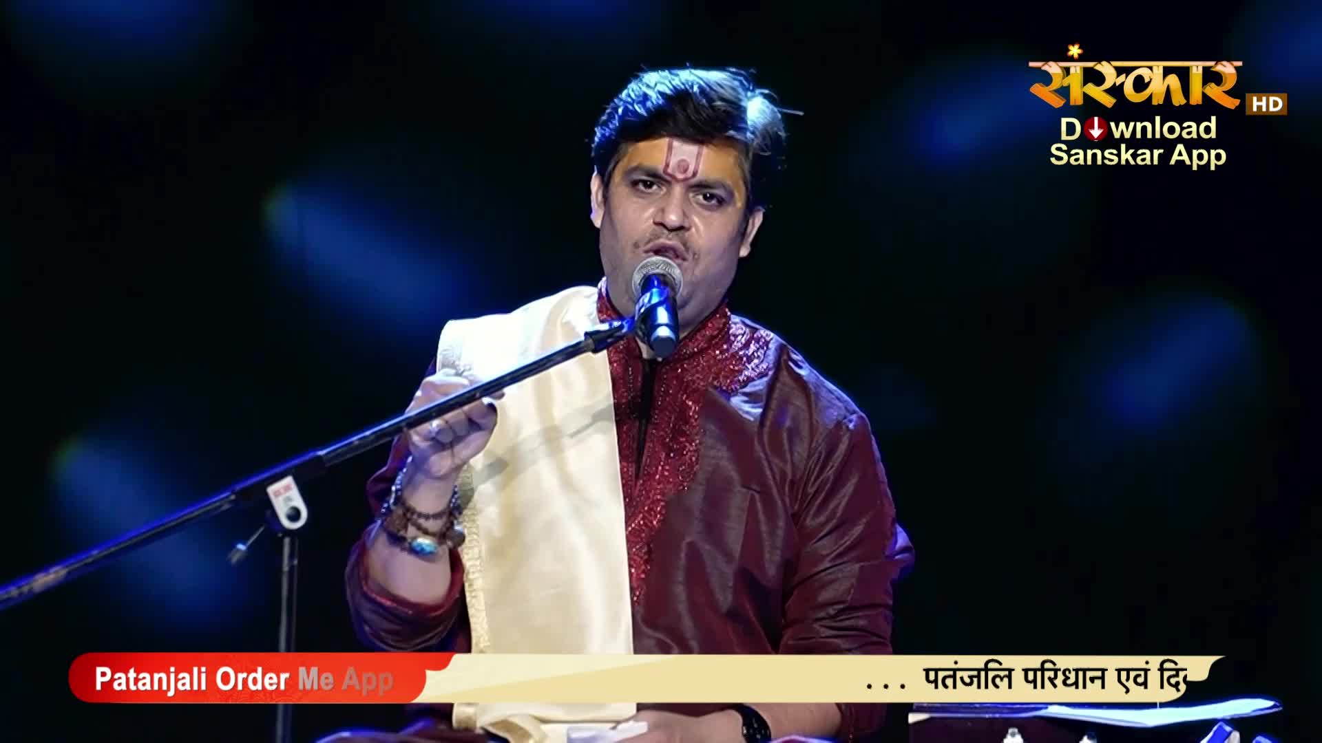 A man in a rich maroon and gold outfit sings into a microphone, his right hand gesturing. The Sanskar TV logo is visible in the upper right corner.