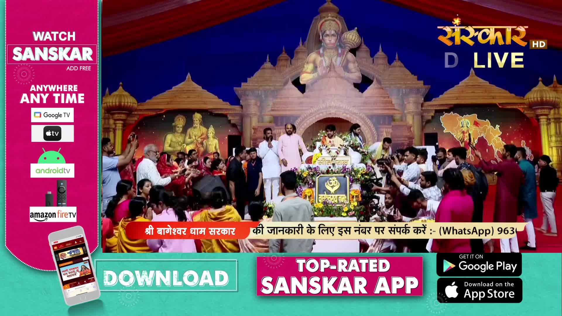 A man in a light pink kurta stands on a raised platform, speaking into a microphone. A crowd of people, many holding up their phones, surround the stage, their faces turned towards the speaker. The backdrop features a large, ornate structure with statues of deities.