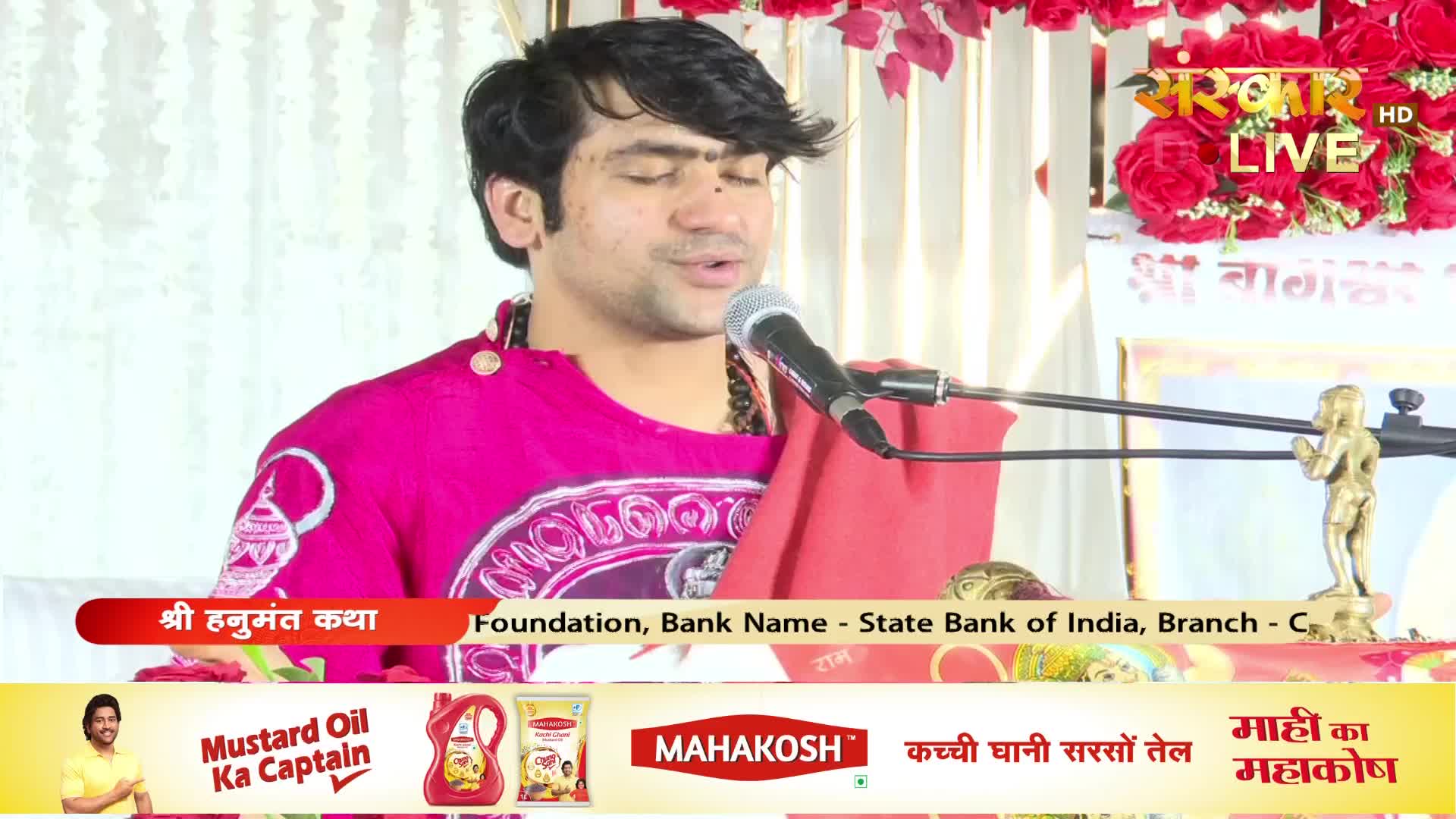 A man in a bright pink kurta speaks into a microphone, his eyes closed. Behind him, a banner reads "Sanskar Live" and a small golden statue of a deity stands on a stand.