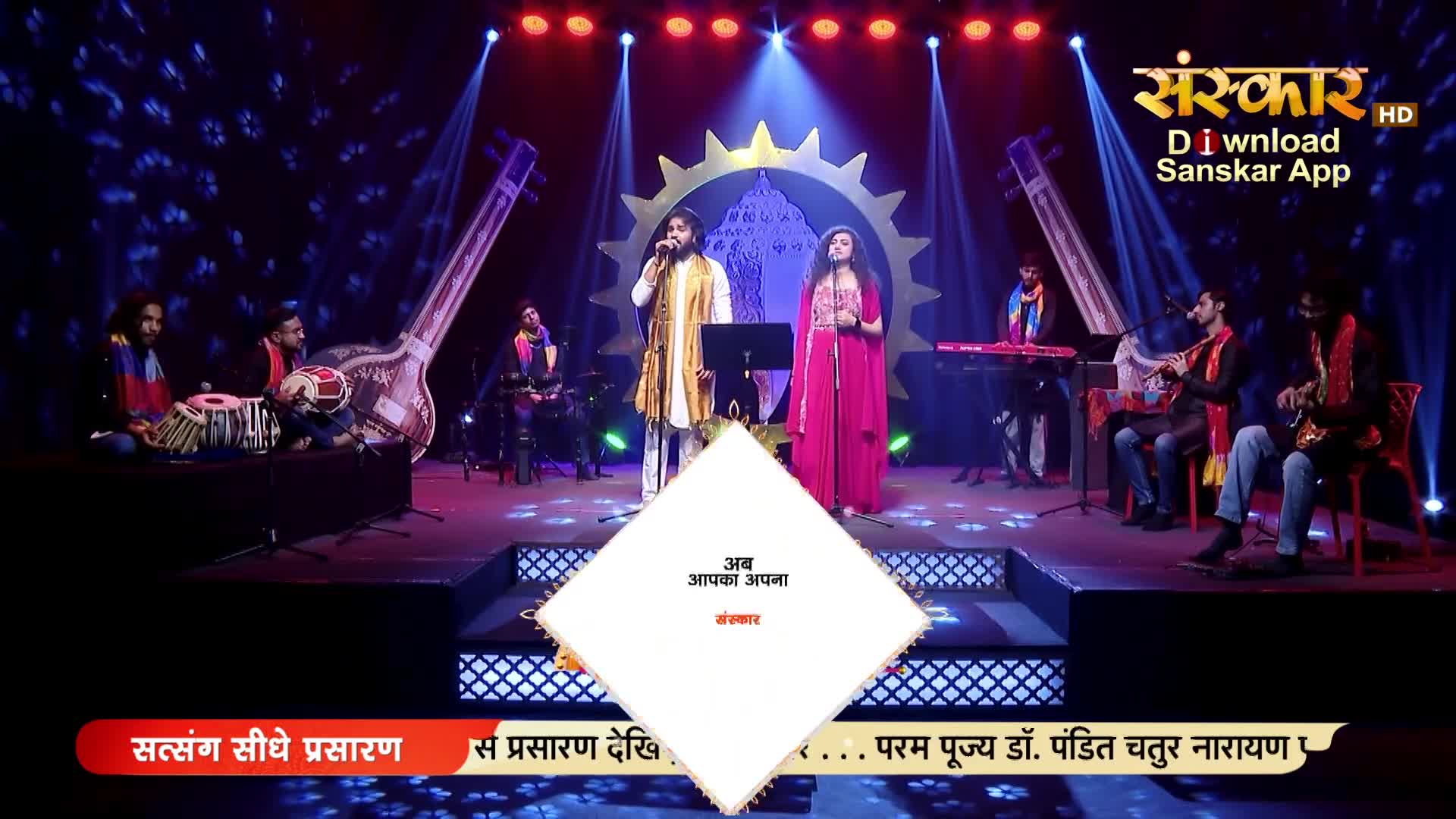 A male and female singer stand center stage, bathed in light. Musicians play traditional Indian instruments around them, their melodies filling the air.