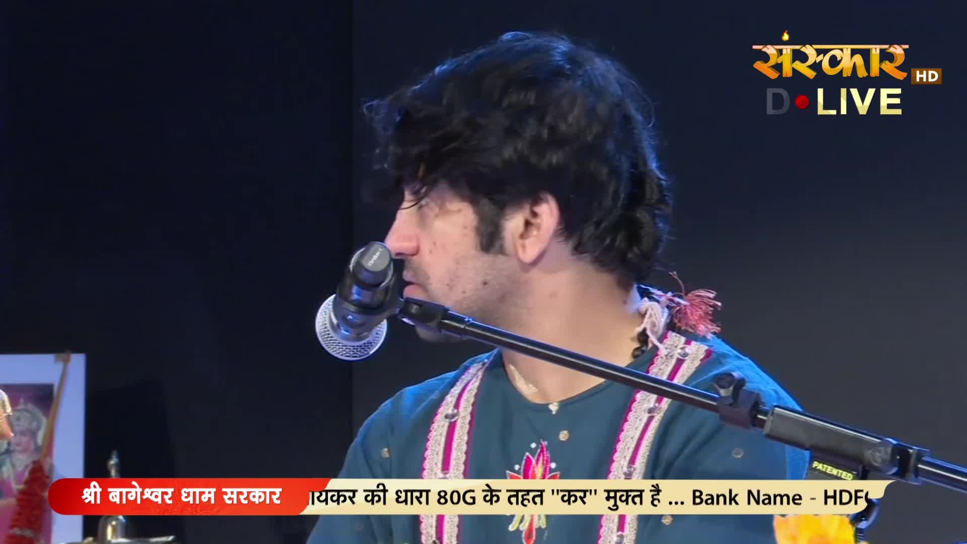 A man with dark, wavy hair leans into a microphone, his profile sharp against the dark background. He wears a teal kurta with intricate pink and white embroidery, a traditional Indian garment.