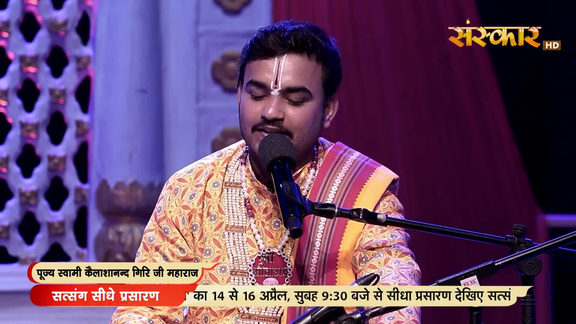 A man in a vibrant yellow and orange kurta, adorned with a beaded necklace, sings into a microphone. The Sanskar HD logo and text indicating a live Satsang broadcast are visible.