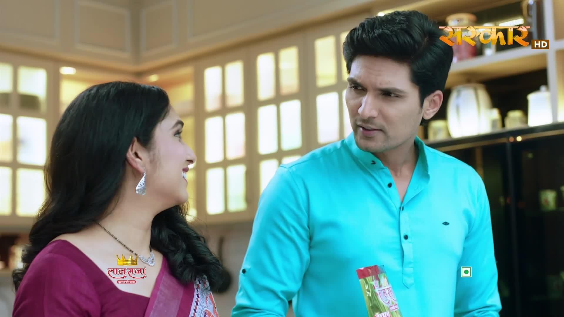 A man in a teal kurta holds a box of what looks like snacks, turning to speak with a woman in a magenta sari. The Sanskar TV logo is visible in the background.