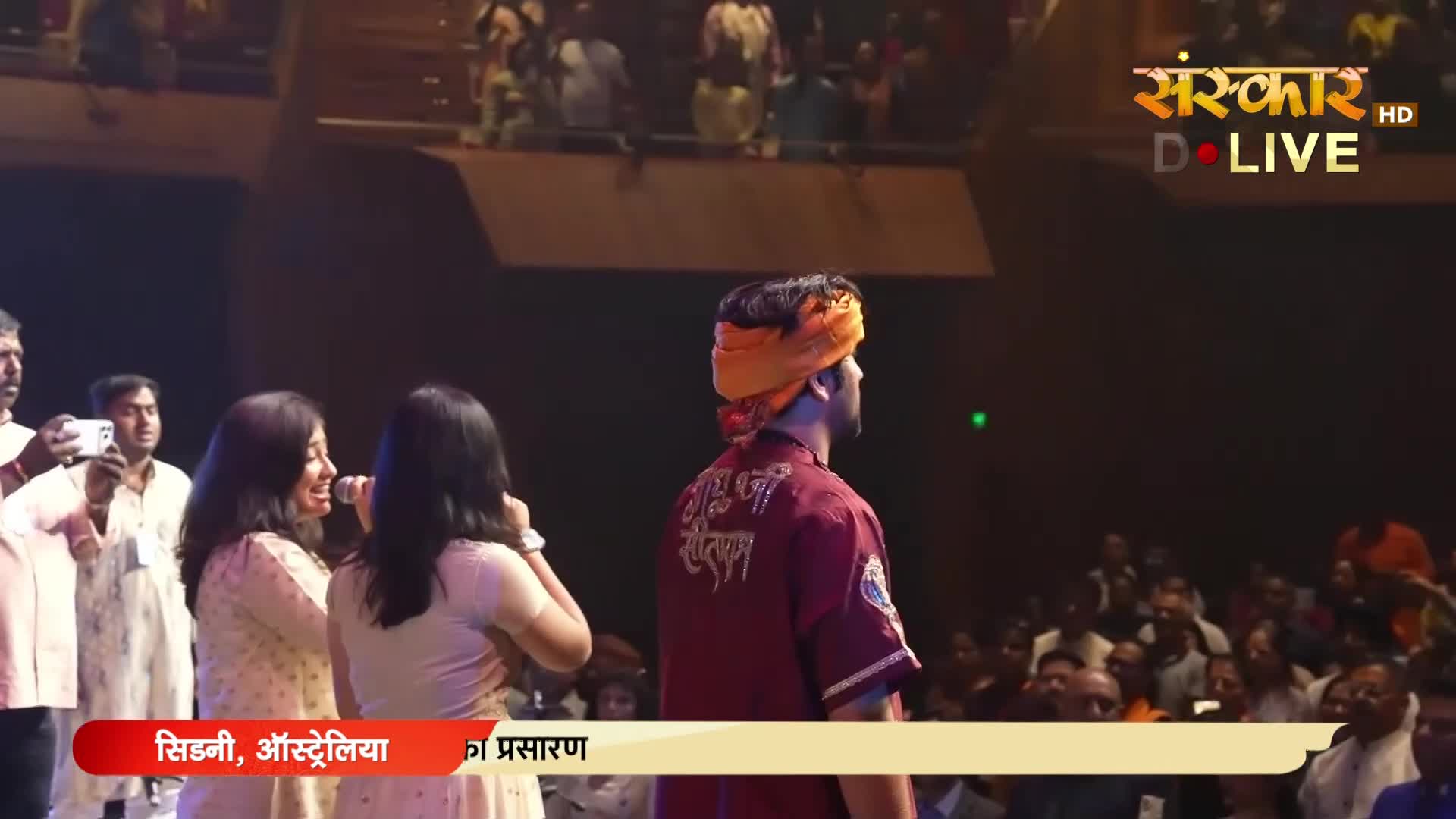 A man in a maroon uniform with an orange turban stands on a stage, facing away from the audience. Two women in light-colored dresses are to his left, one holding a microphone.