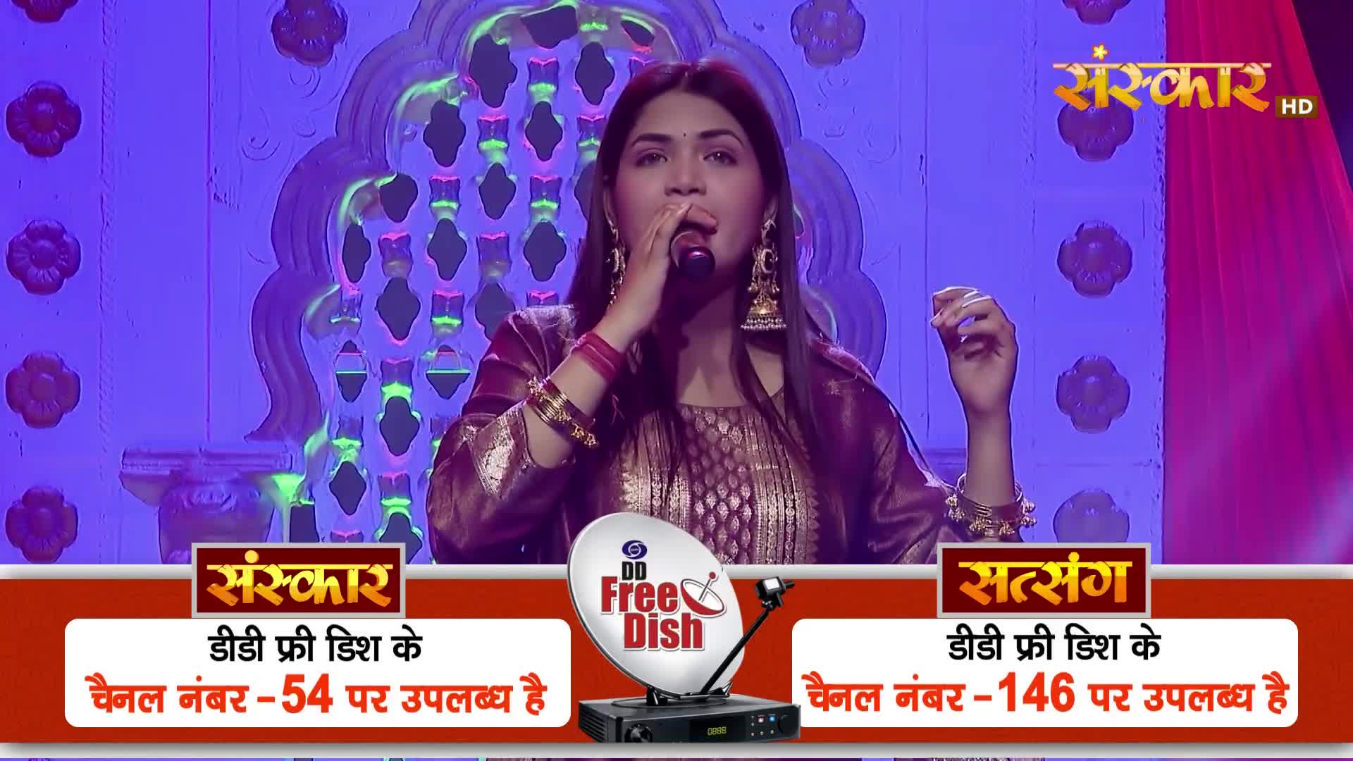 A woman sings into a microphone on a stage, her voice echoing. Below her, a satellite dish and receiver sit on a red and gold platform.