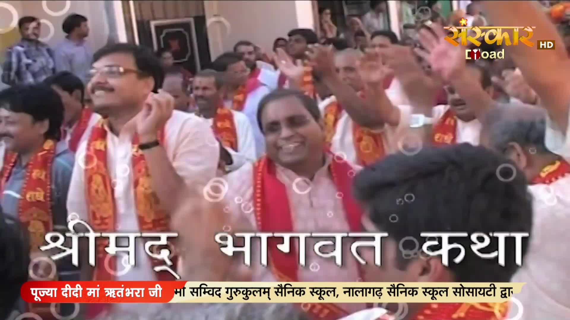 A crowd of men in white kurtas and red sashes are cheering, their hands raised in celebration. The Sanskar TV logo is visible, suggesting a broadcast of a religious event.