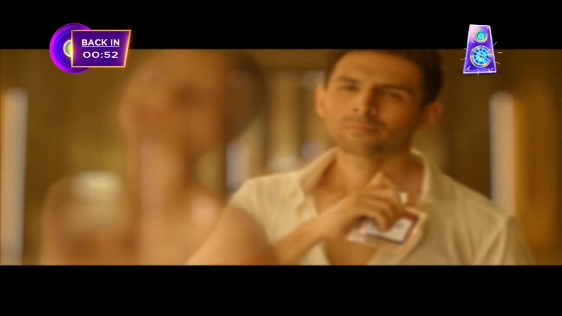 A man in a white shirt holds a small card, looking down at it thoughtfully. Behind him, a woman is out of focus, and a timer on the screen shows the show is "back in" under a minute.
