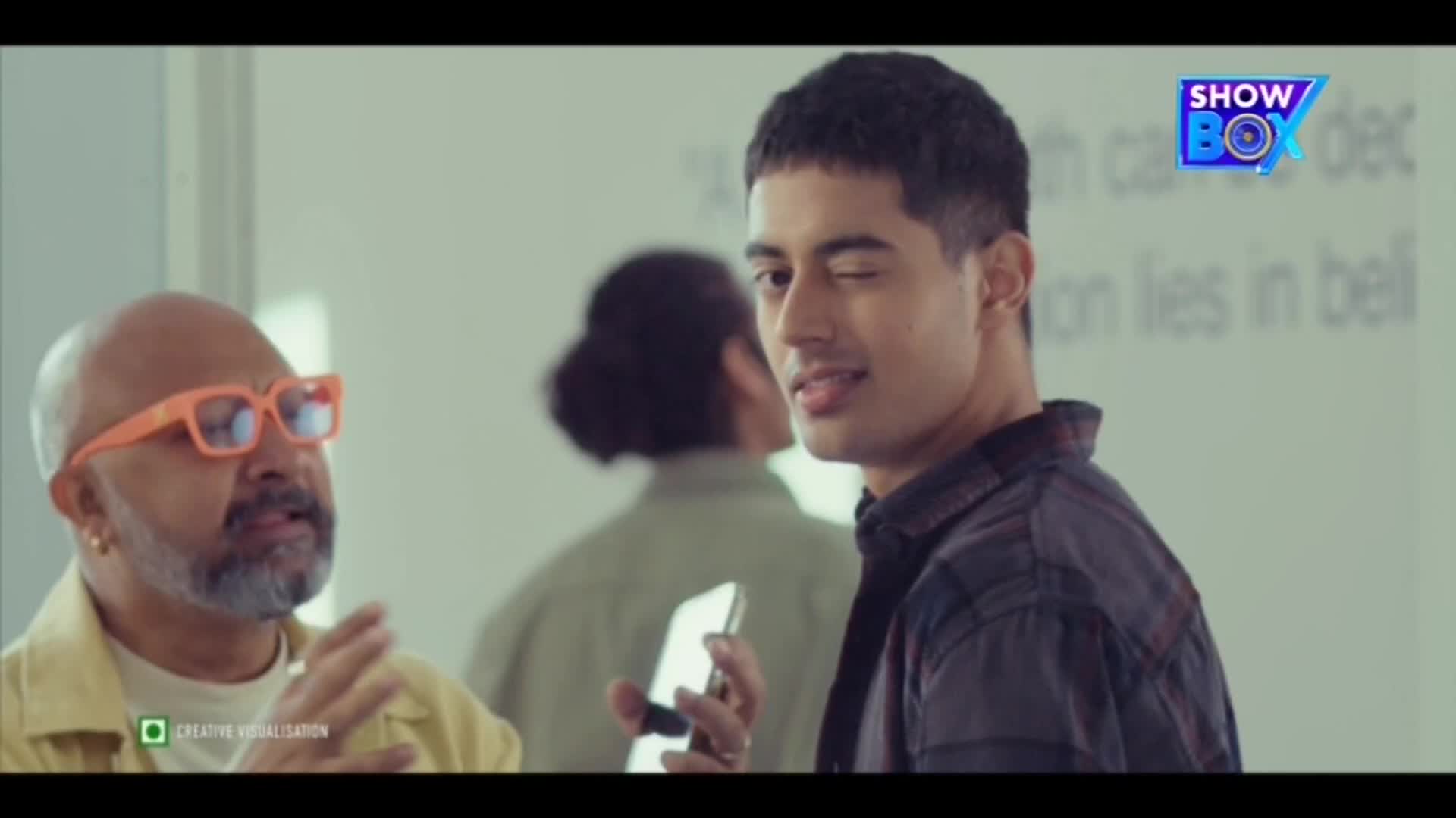 The man with the orange glasses is talking, gesturing with his hands. A younger man with a phone in his hand winks, looking directly at the camera.
