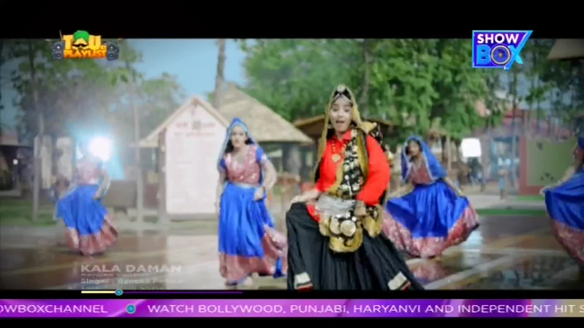 Dancers in vibrant outfits are moving in a choreographed routine, their skirts swirling as they step. The scene is from a music video, likely broadcast on ShowBox, with a backdrop of what looks like a village setting.
