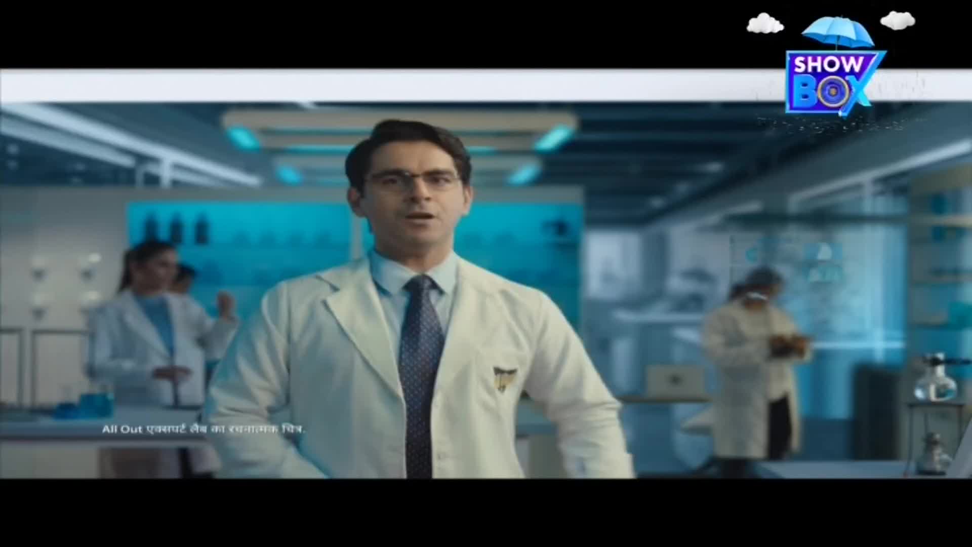 A man in a lab coat is speaking directly to the camera, while a ShowBox logo hovers above. Behind him, lab assistants work in a sterile environment.
