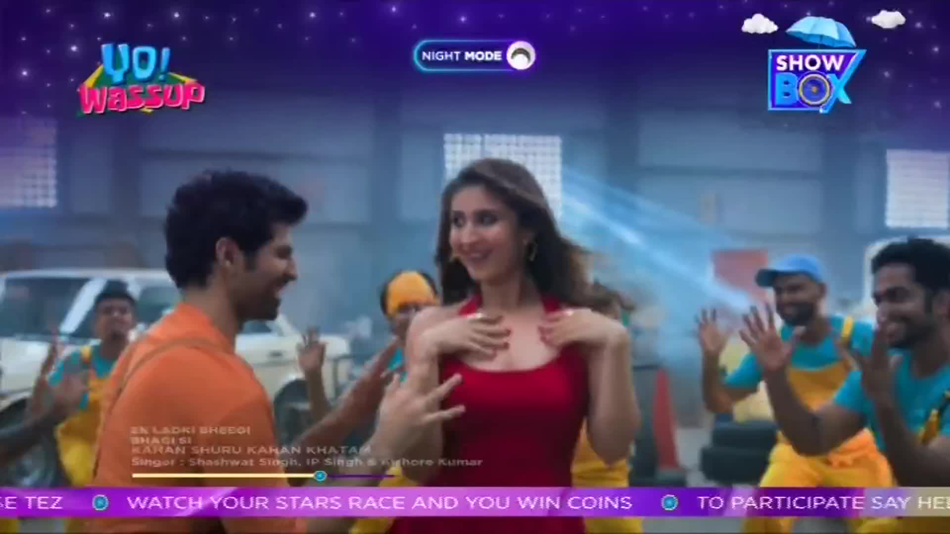 A woman in a red dress and a man in orange smile and dance, surrounded by a group of men in blue and yellow.  The scene is a music video, likely from India, as indicated by the ShowBox logo and the "Yo! Wassup" graphic.
