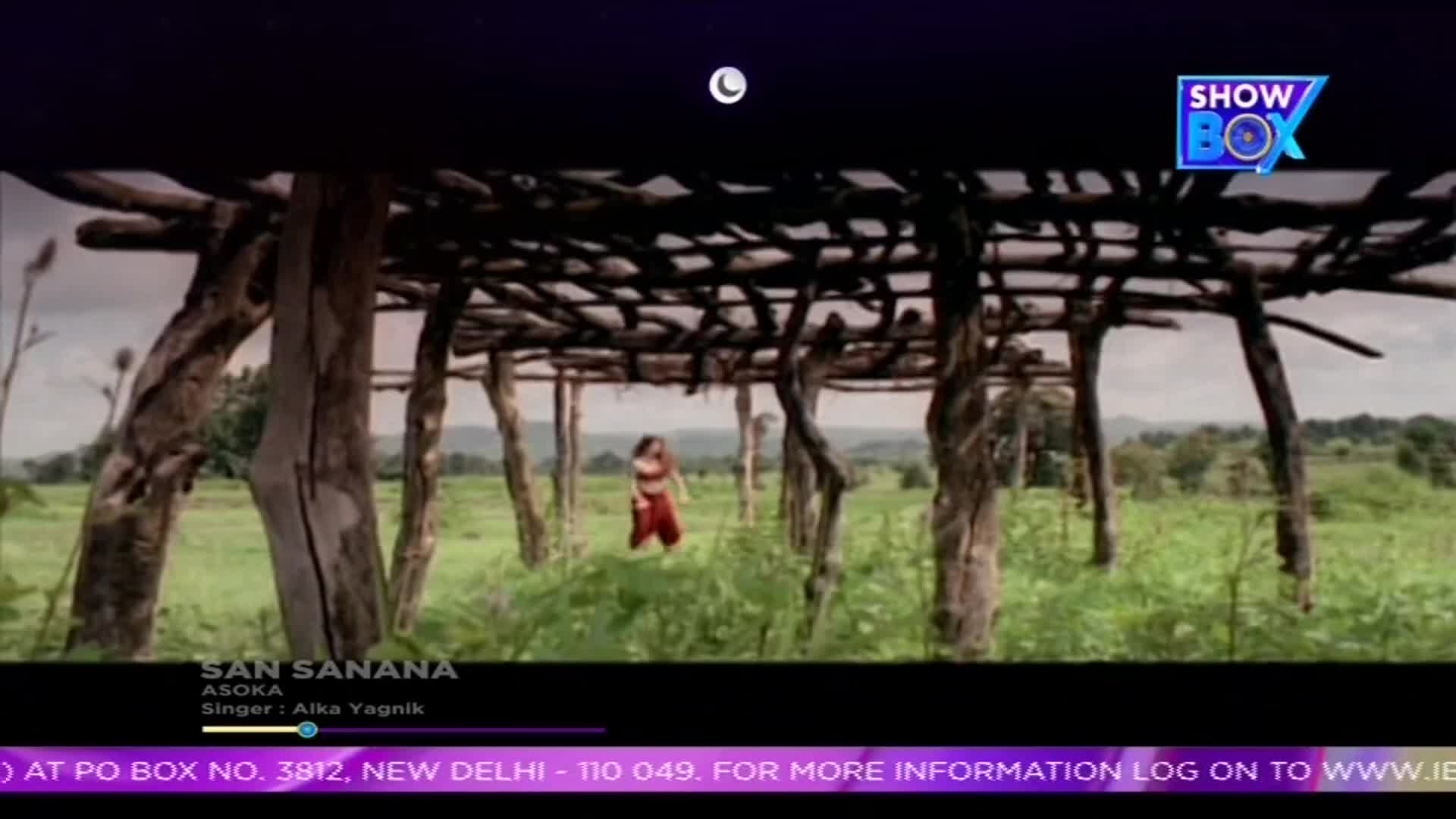 Under a rustic wooden structure, a woman in red and orange clothing moves through tall grass. The scene, broadcast on ShowBox, is set in a field with a backdrop of distant hills.
