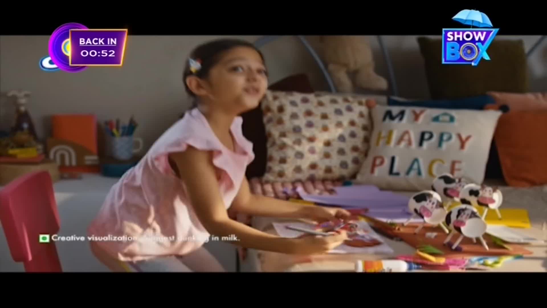 A young girl in a pink dress is seated at a table, working on a drawing.  Cow-shaped crafts are scattered on the table, and the ShowBox logo is visible in the corner.
