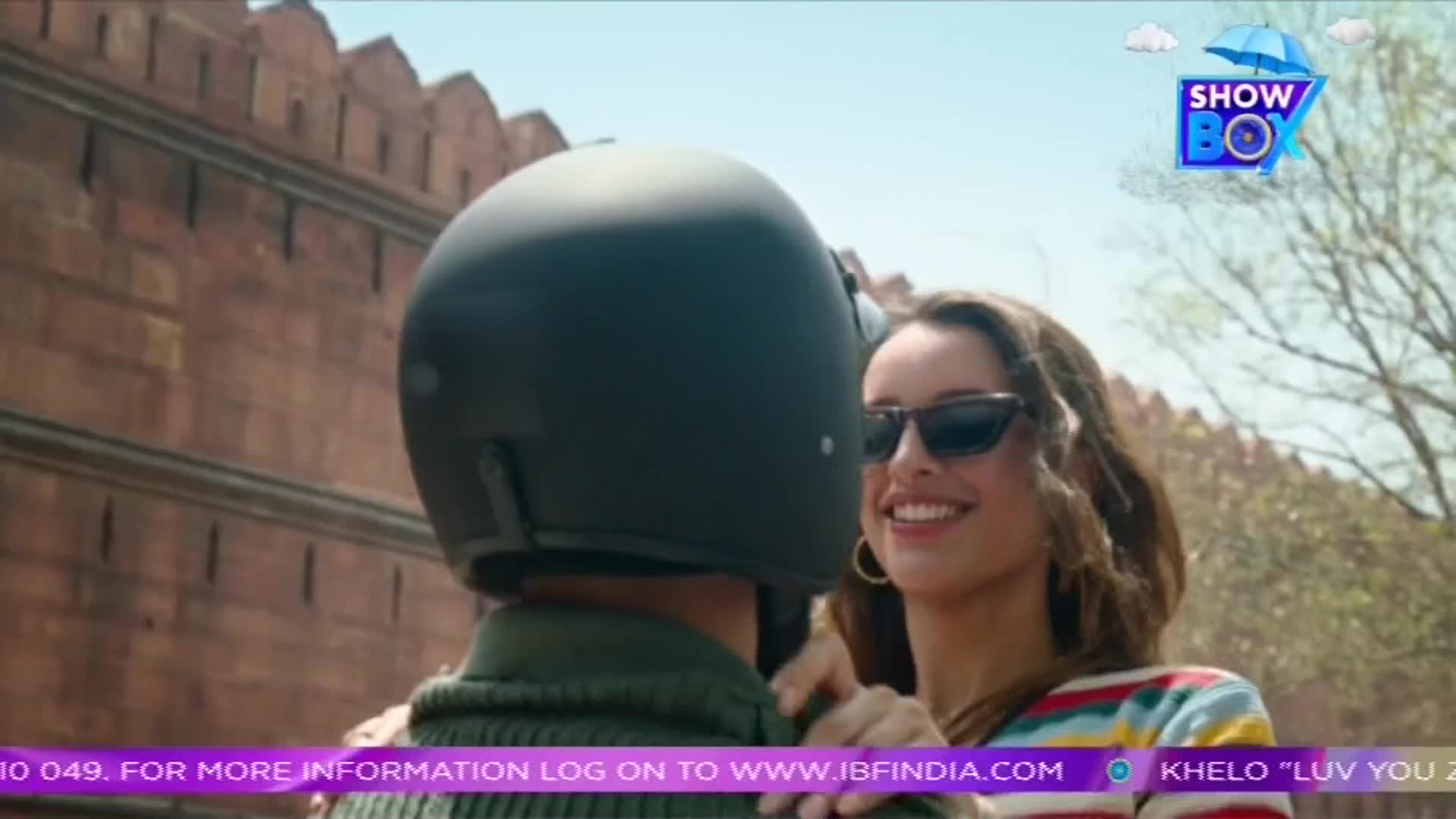 A woman with sunglasses smiles, her arms around a person wearing a black helmet. The scene unfolds in front of a red brick wall, with the ShowBox logo in the corner.
