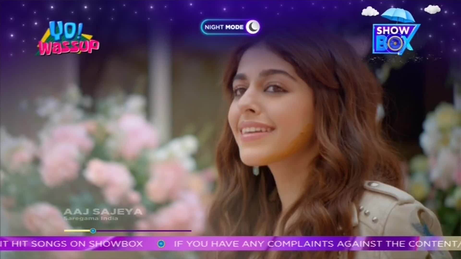 A woman with wavy hair smiles, her gaze directed off-screen. The ShowBox logo and "Aaj Sajeya" text are visible, suggesting a music video or program is playing on a screen.
