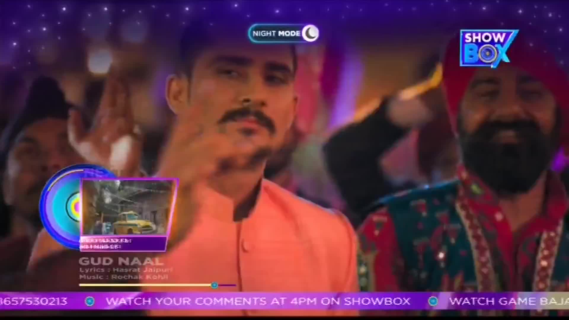 Two men in traditional Indian attire are smiling and looking toward the camera, as a music video plays on ShowBox. A smaller screen shows a clip of a yellow car, while text at the bottom invites viewers to watch comments.
