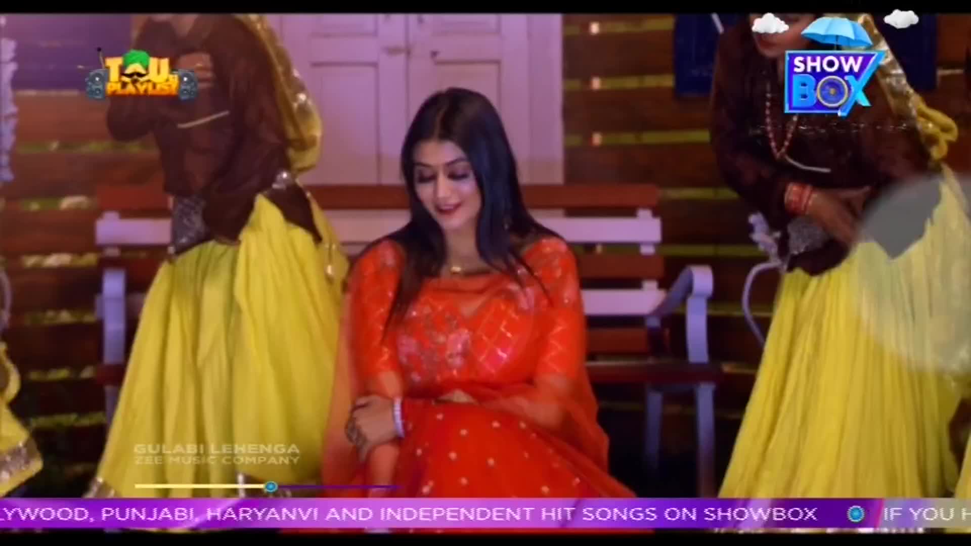 A woman in a bright red outfit sits on a bench, smiling, as dancers in yellow skirts and brown tops move behind her. The scene appears to be from a music video, possibly broadcast on ShowBox, with text at the bottom advertising Indian music.

