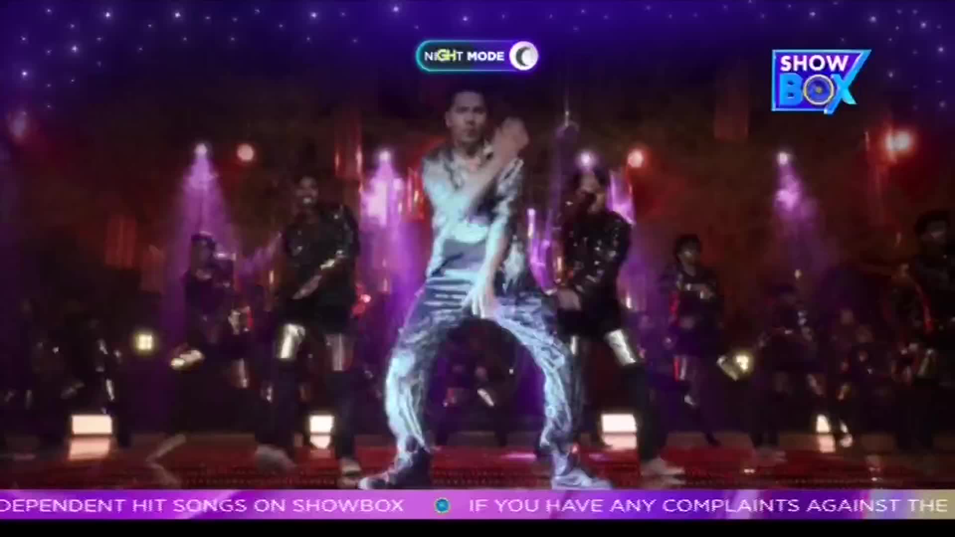 A performer in a shimmering outfit is center stage, striking a pose with his legs wide. Behind him, other dancers in black outfits move in sync, illuminated by purple stage lights.
