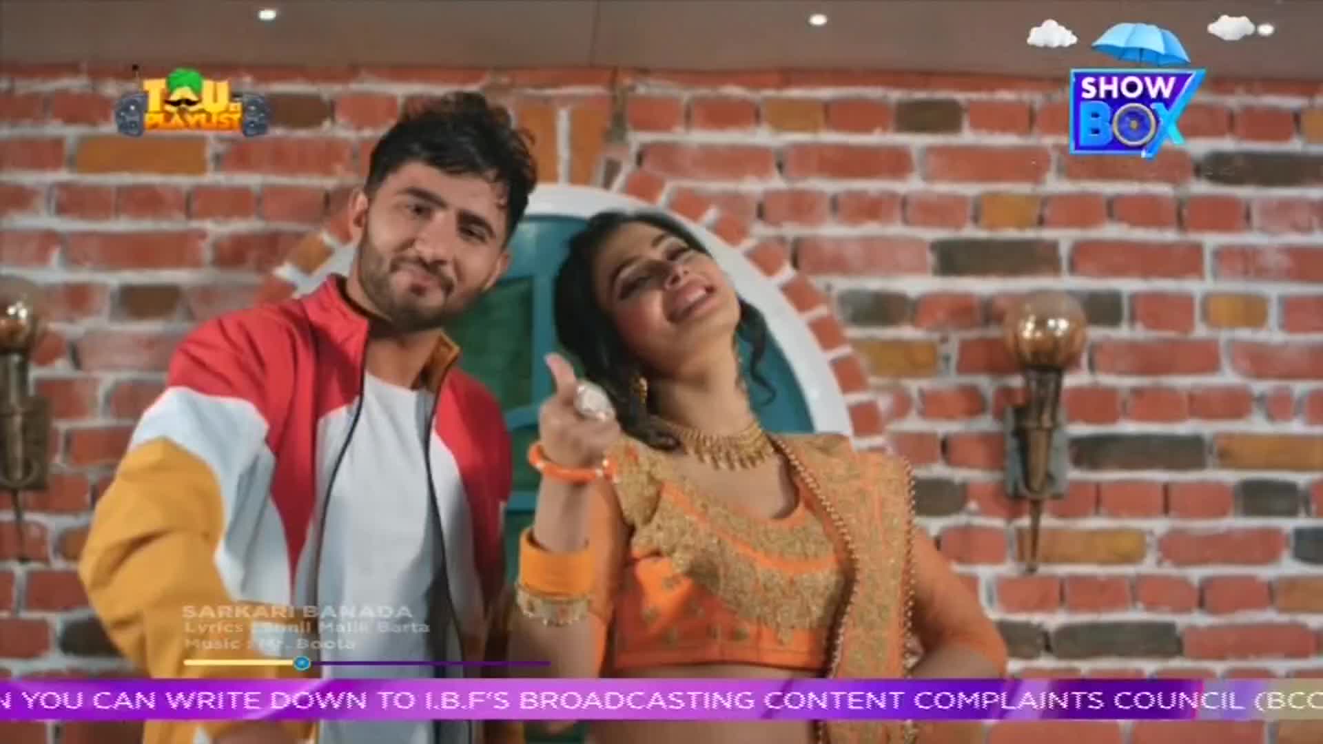 A man in a red and white jacket stands next to a woman in a gold sari, both smiling. The ShowBox logo is visible in the top right corner, suggesting this is a music video from India.
