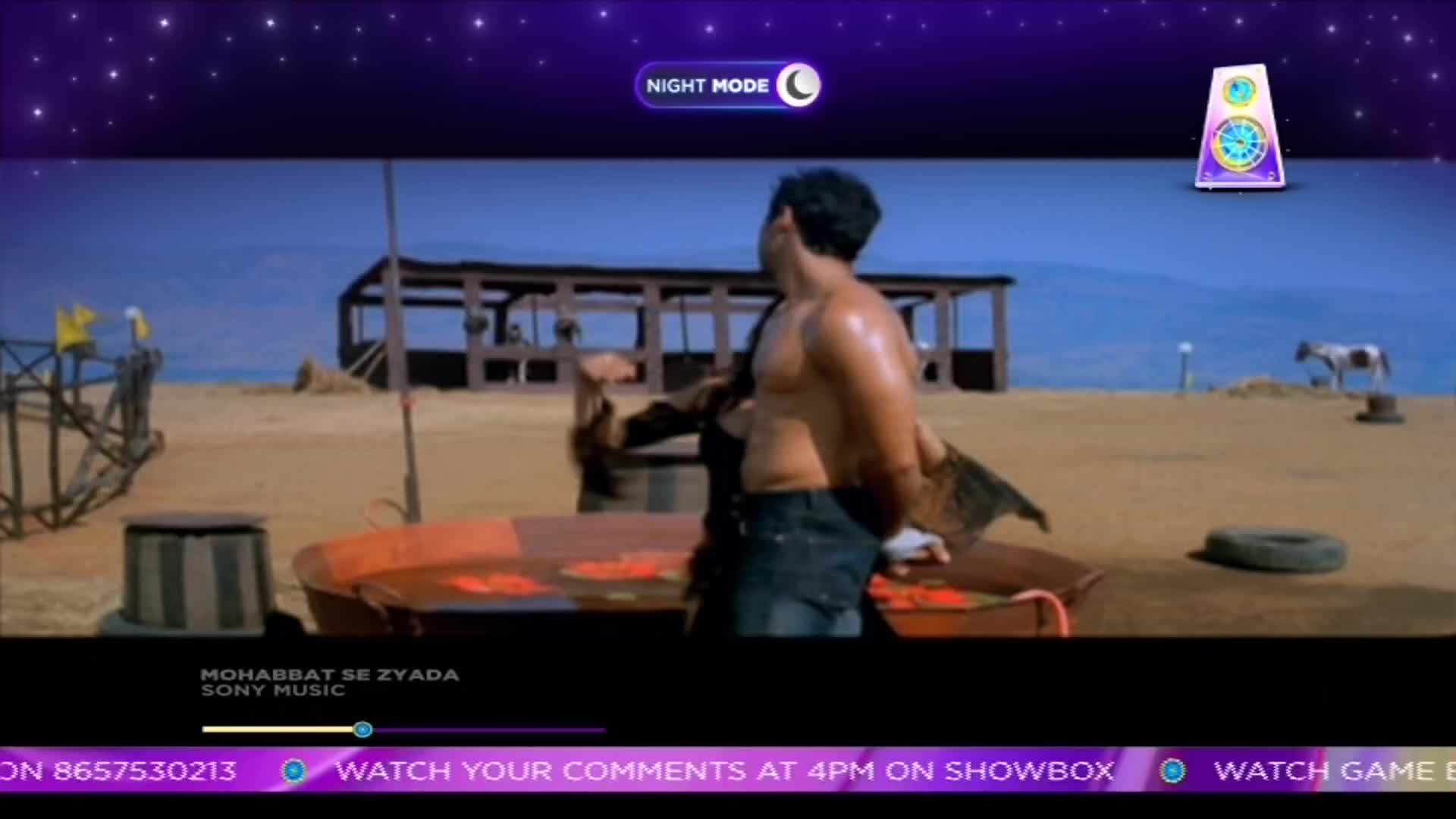 A shirtless man, glistening with sweat, gestures towards someone off-screen. The scene unfolds on a sandy expanse, likely a music video from India, with a ShowBox promotion running at the bottom.
