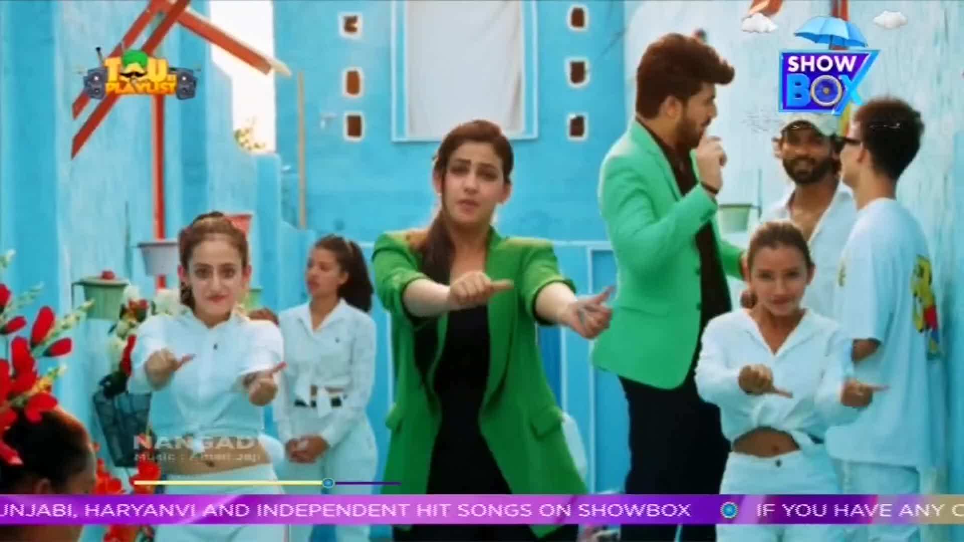 A woman in a green blazer and a black top gestures with her hands, flanked by dancers in white outfits. A man in a green jacket holds a microphone, as the scene unfolds in front of a blue backdrop, likely for a ShowBox music video.
