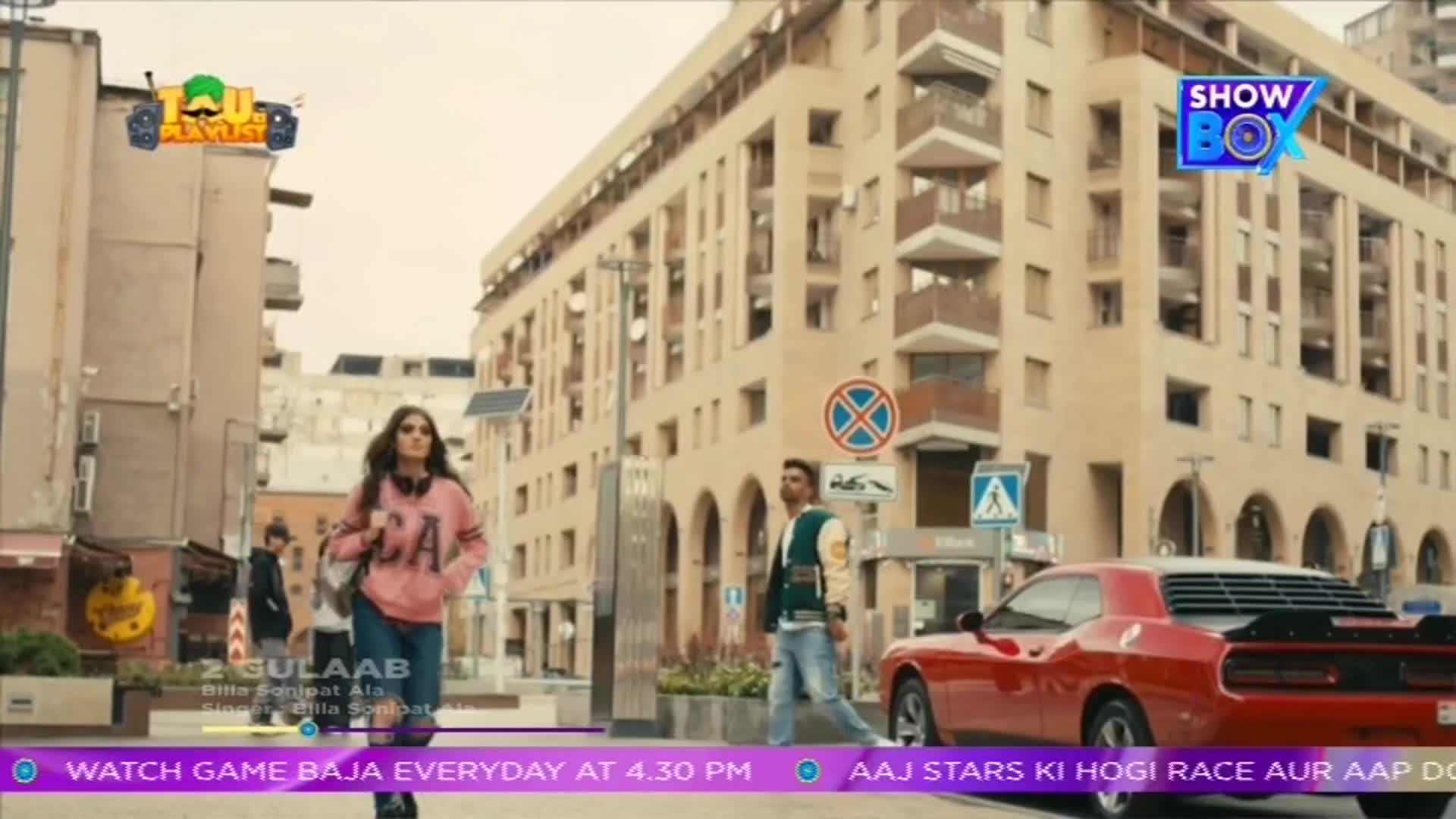 A woman in a pink sweatshirt strides toward the camera, while a man in a letterman jacket crosses the street near a bright red car. The scene unfolds on a city street, likely part of a music video from ShowBox in India.
