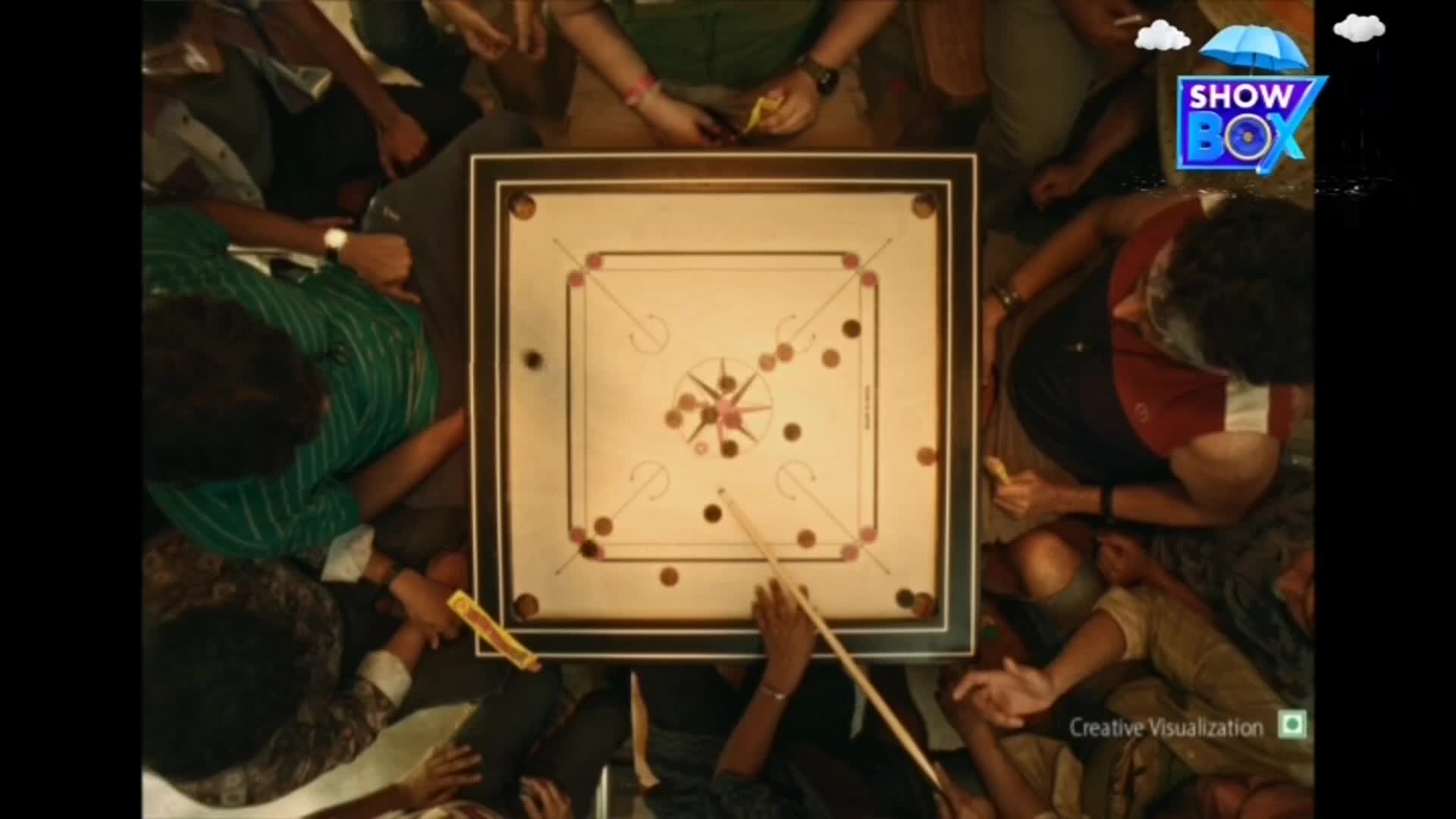 A game of carrom is underway, with players gathered closely around the board. The striker is poised, aiming to send a disc flying into a pocket.
