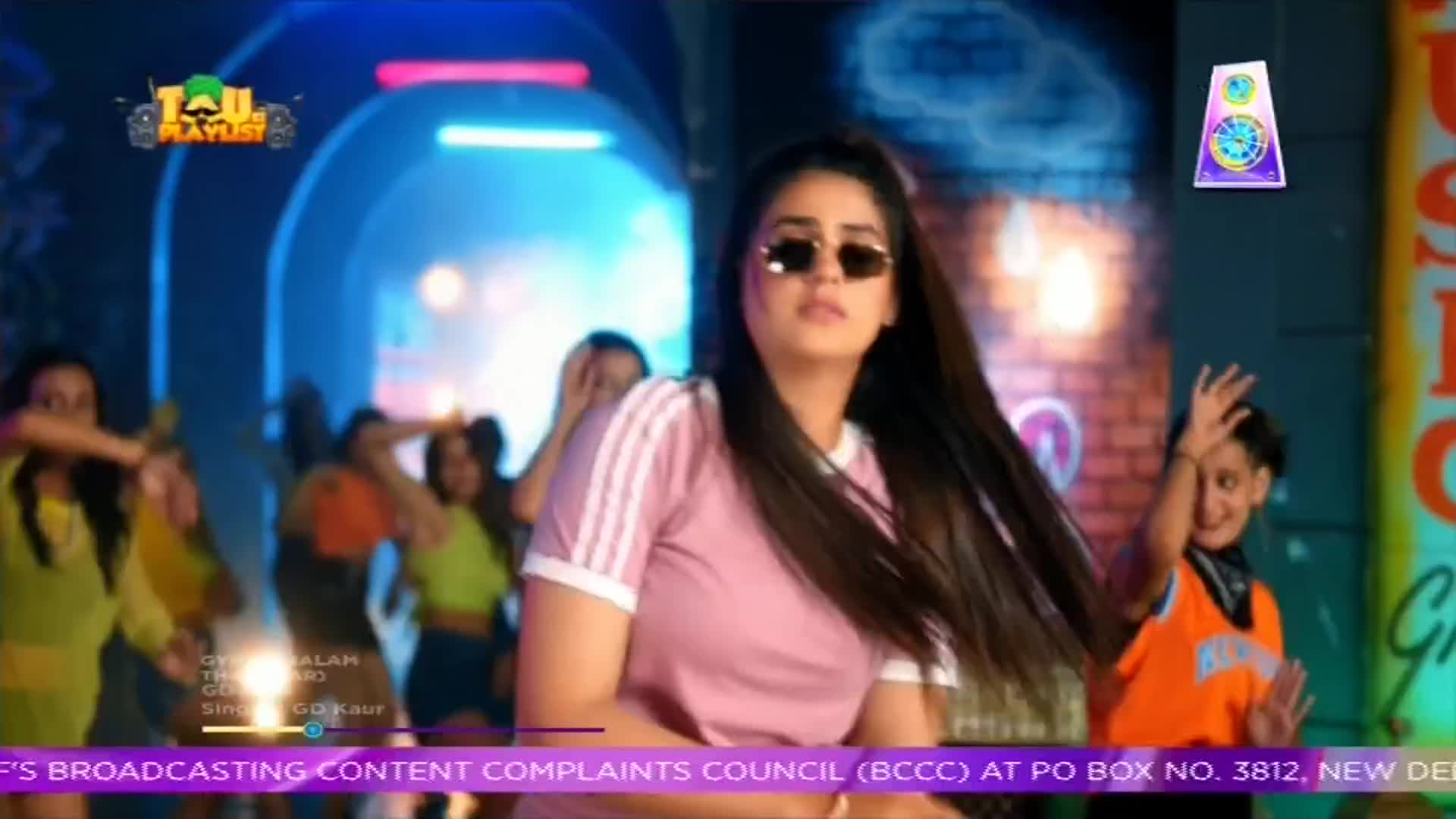A woman in sunglasses and a pink top is dancing, her long, dark hair flowing. Behind her, a group of people move in time to the music, part of a music video from India, possibly on ShowBox.
