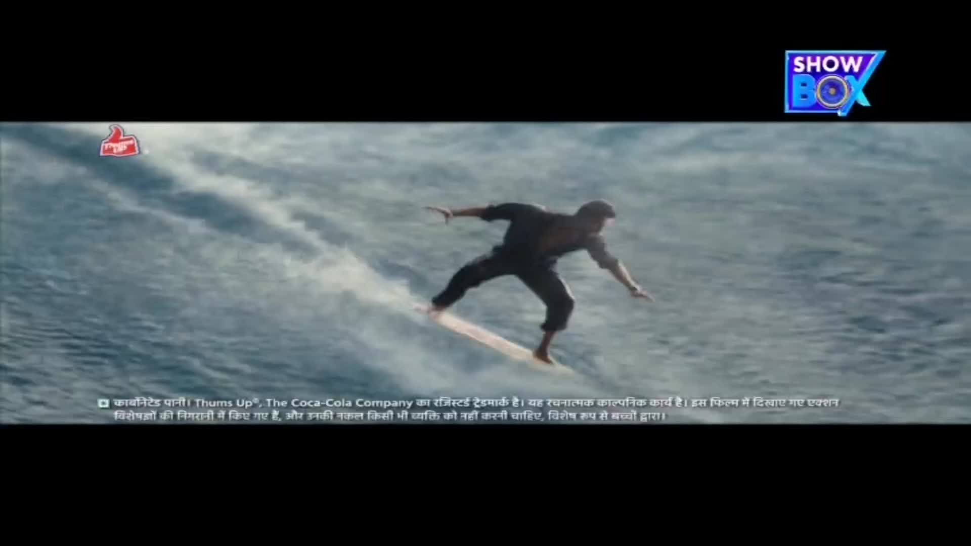 A man in dark clothing balances on a surfboard, carving a path through the ocean. A ShowBox logo and a Thums Up logo are visible, suggesting this is an advertisement from India.
