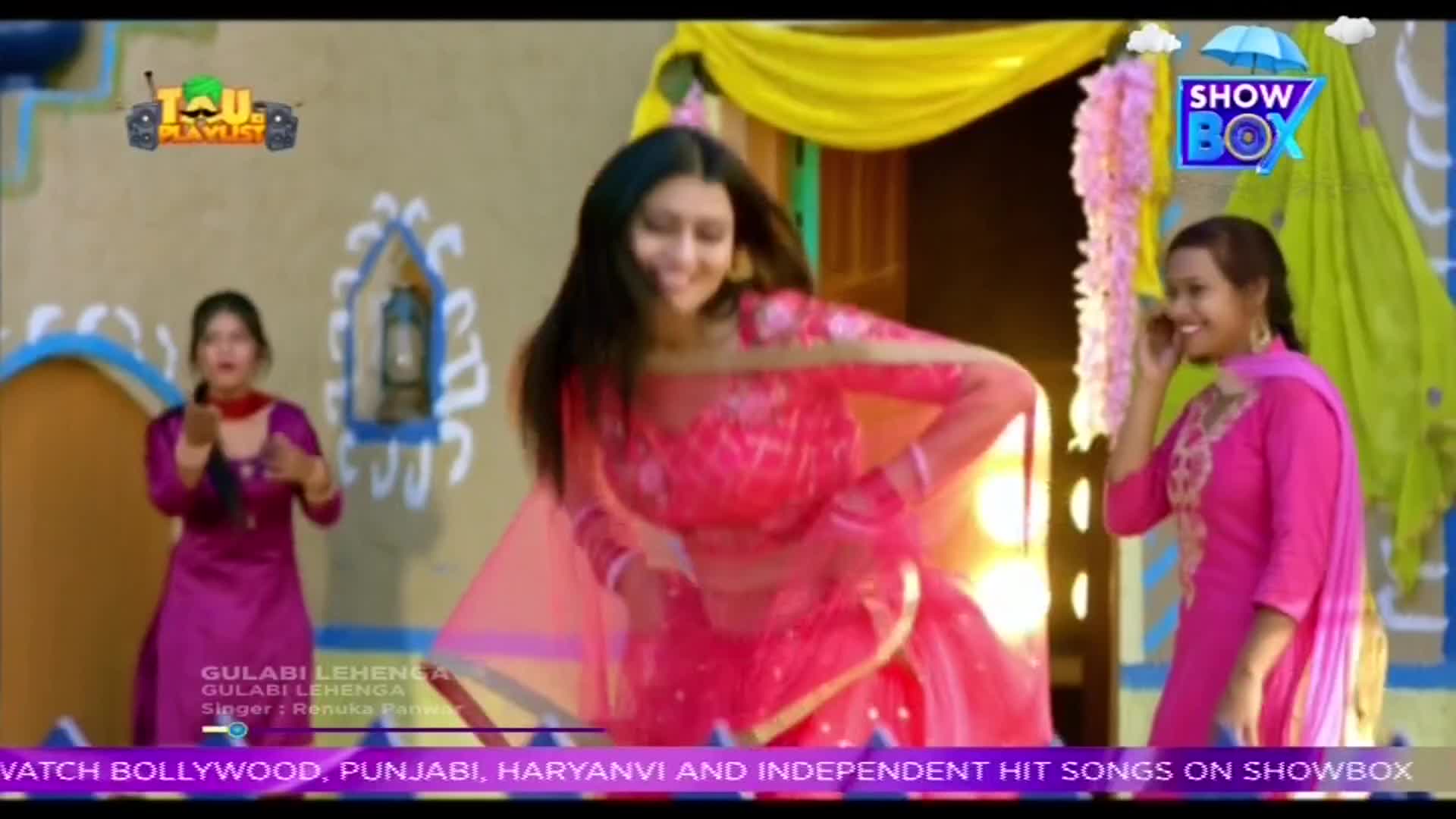 A woman in a bright pink outfit dances in front of a painted backdrop, while two other women in similar attire stand nearby. The scene looks like it's part of a music video from India, possibly on ShowBox.
