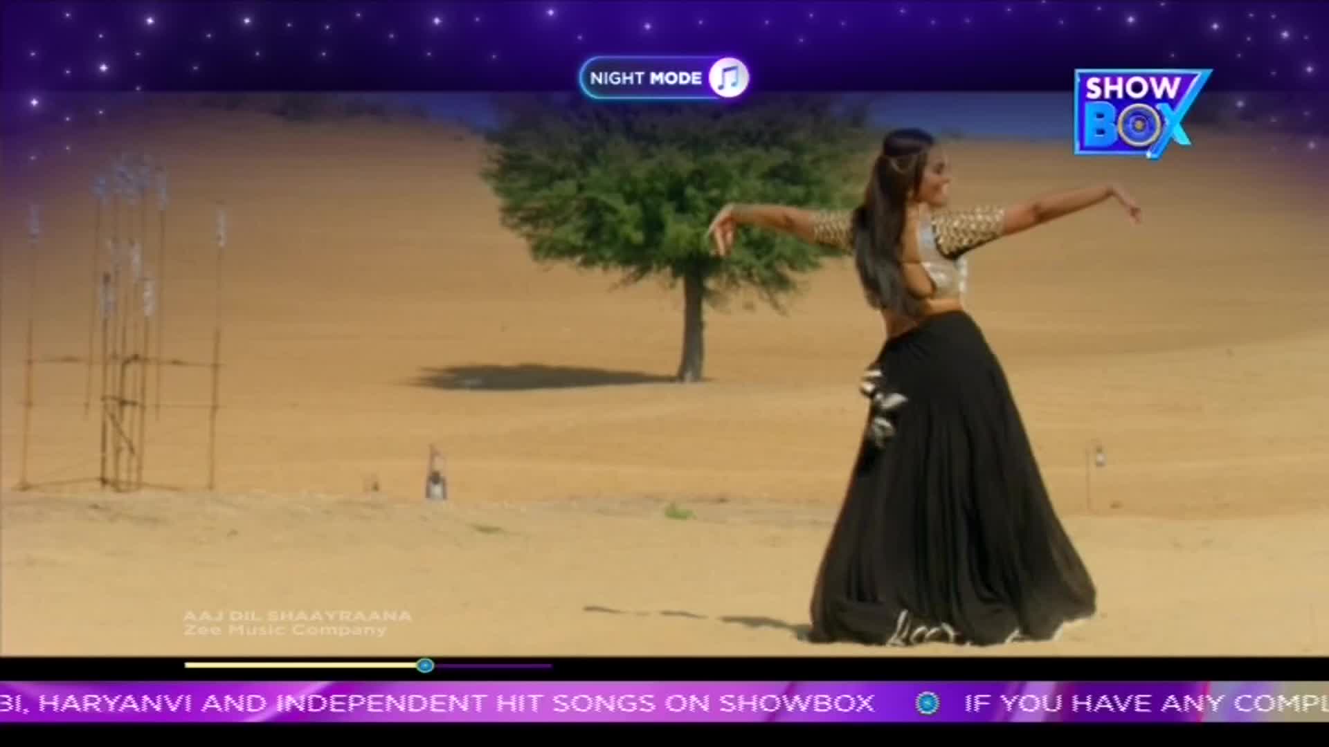 A woman in a black skirt and silver top is dancing in the desert, her arms outstretched. The ShowBox logo is visible, and the screen shows a music video from India.
