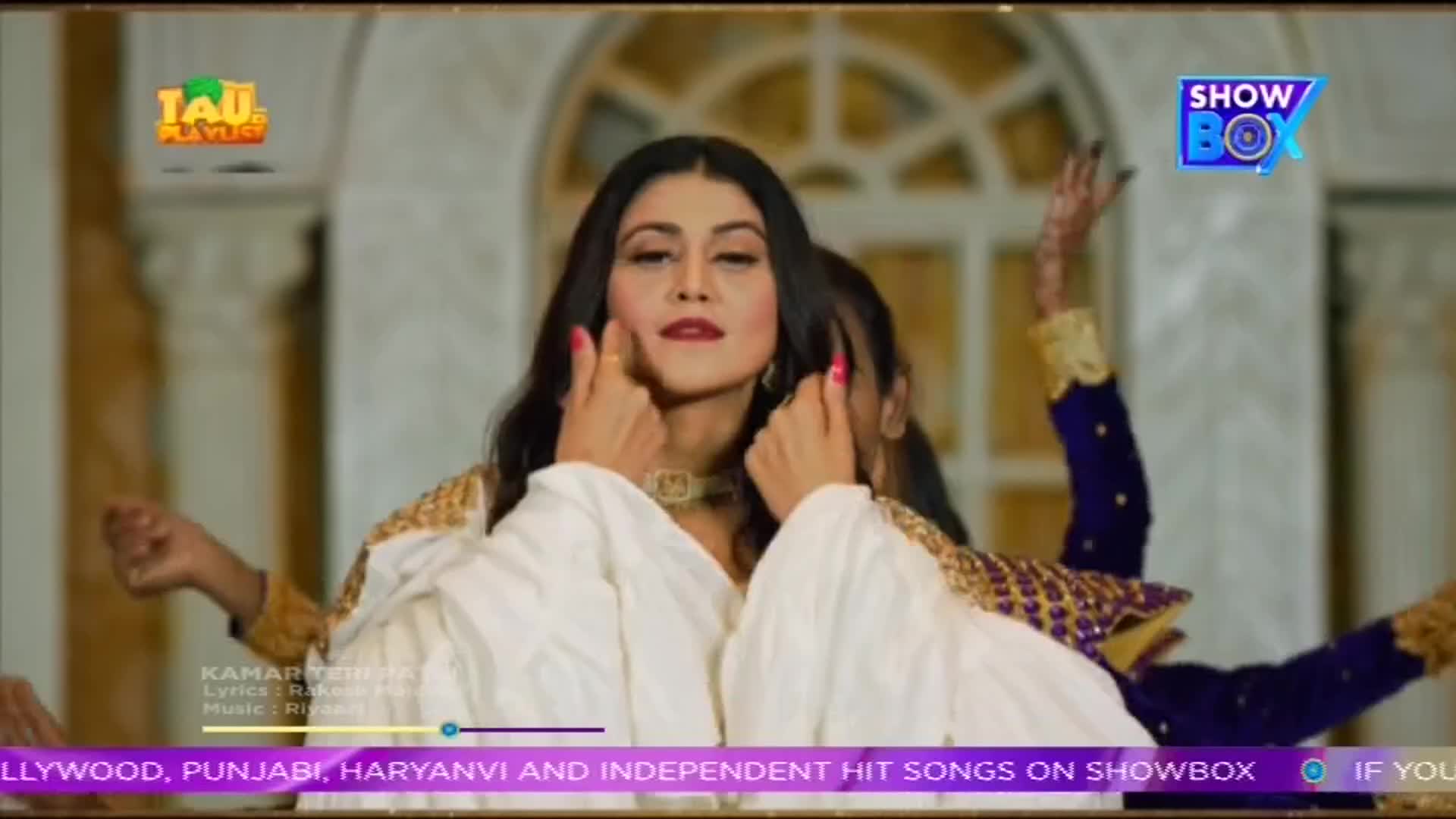 A woman in a white outfit gestures with her fingers, her face framed by long, dark hair. Other figures with raised arms, wearing gold and navy, surround her in what looks like a music video on ShowBox.
