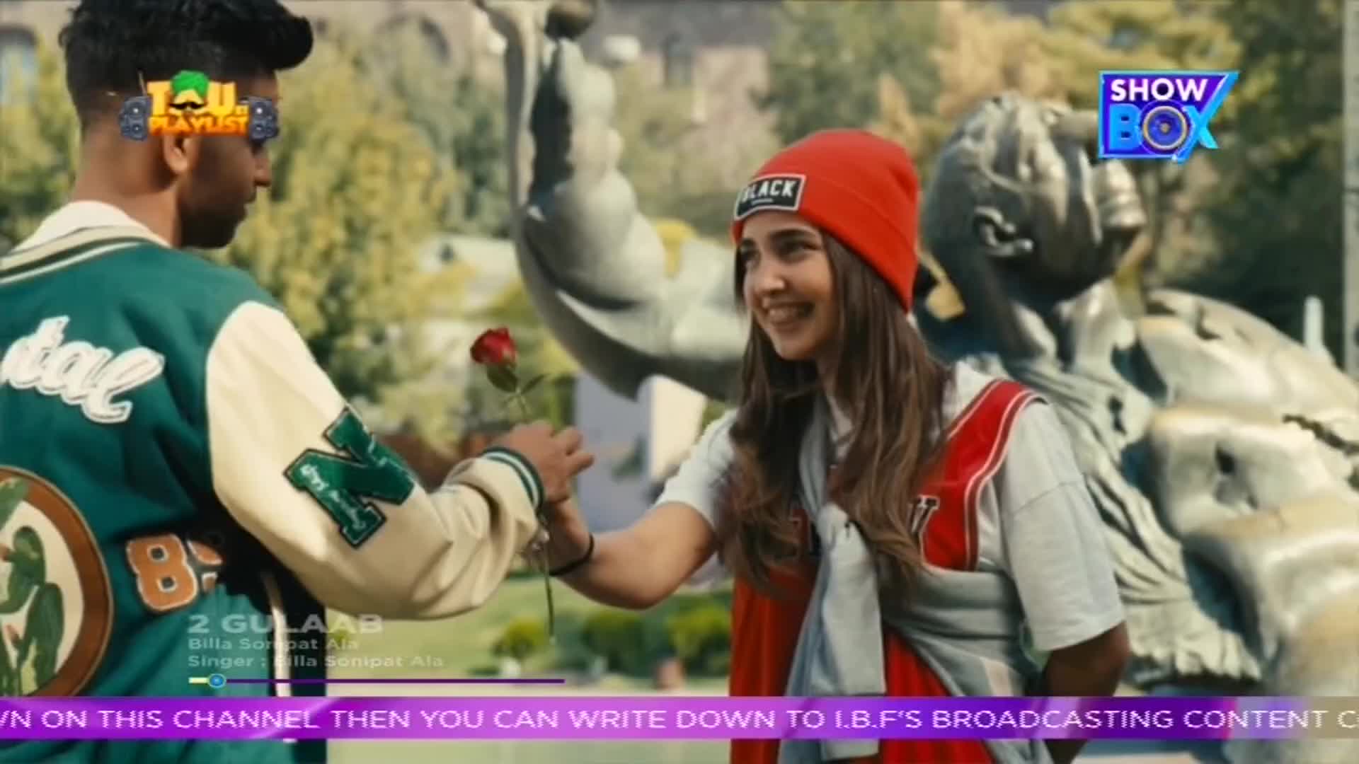 A young man in a varsity jacket offers a red rose to a smiling woman wearing a red beanie. The scene unfolds in front of a statue, likely from a music video broadcast on ShowBox.
