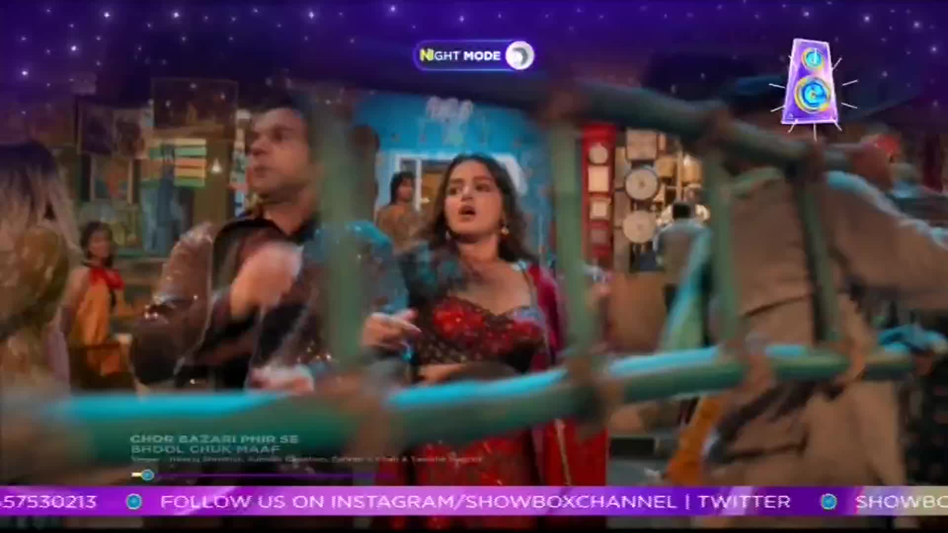 A man with a sparkly jacket and a woman in a red outfit are caught in the middle of the action, looking surprised. Someone is carrying a long, blue-painted ladder, obscuring much of the scene on this ShowBox broadcast.
