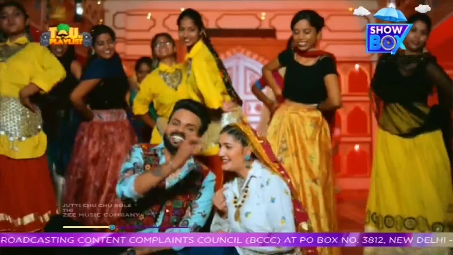 A group of women in colorful skirts and tops are dancing, with some clapping their hands. A man in a patterned shirt laughs, gesturing towards a woman in a white top and headscarf.
