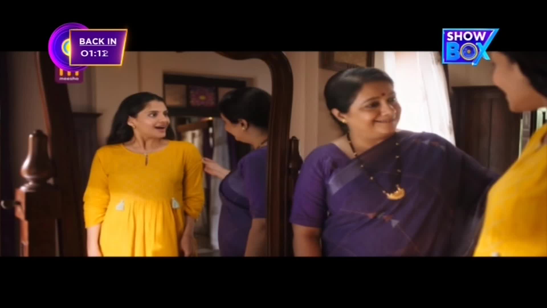 A woman in a yellow dress looks at herself in the mirror, while another woman in a purple sari smiles at her. The scene is part of a ShowBox program, with a "Back in 01:12" graphic in the corner.
