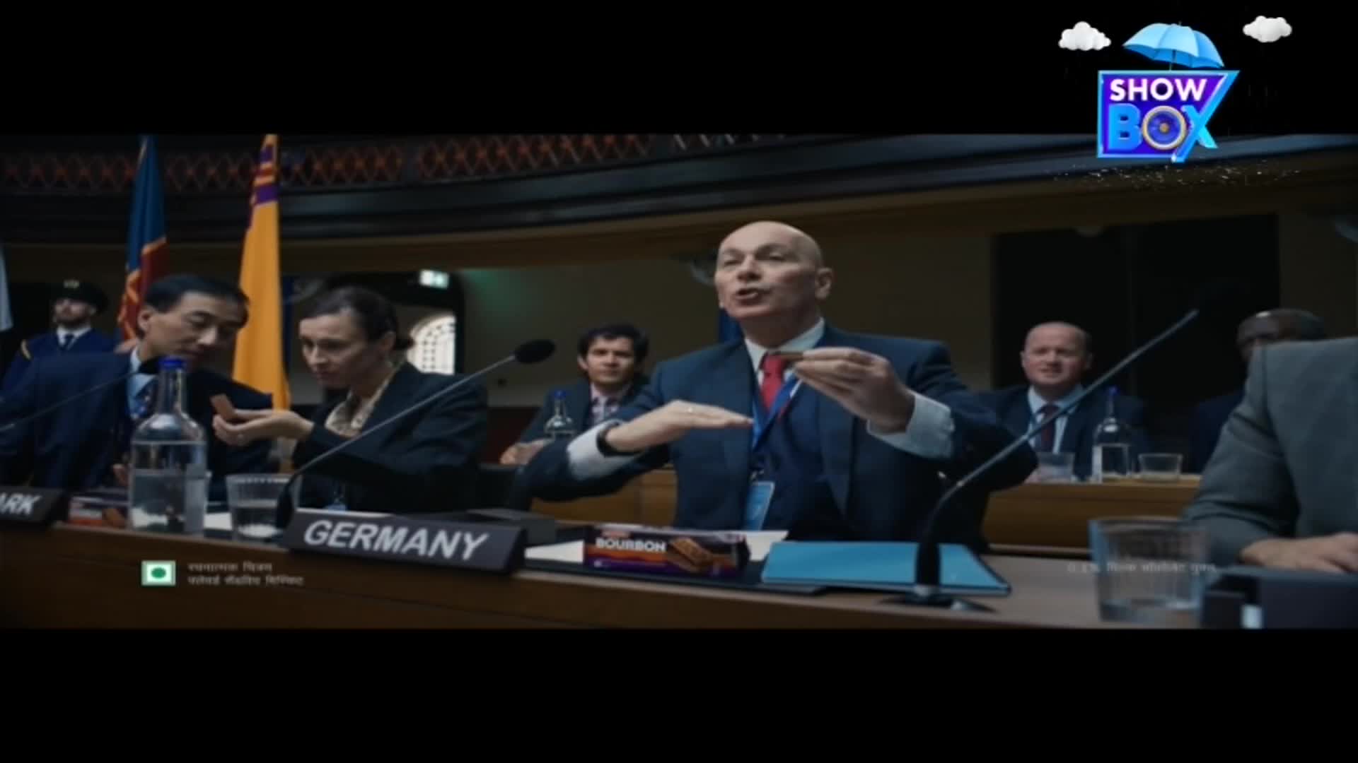 A man in a blue suit gestures with a chocolate biscuit, speaking with animated expression.  The scene is set at a conference table, with the "ShowBox" logo in the top right corner.
