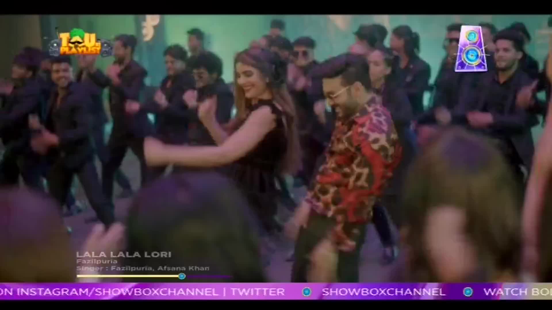 A woman in a black dress and a man in a patterned shirt dance in front of a crowd of people in dark suits. The scene is from a music video on ShowBox, with the song title "Lala Lala Lori" displayed on the screen.
