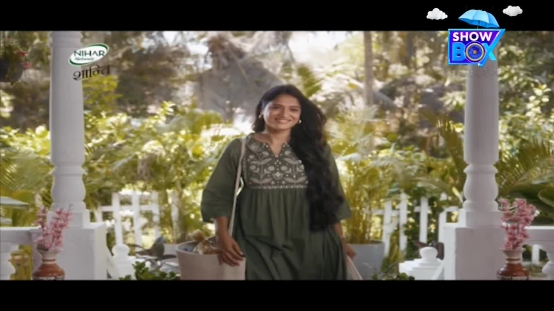 A woman in a green dress smiles as she walks toward the camera, her long black hair flowing. The scene, possibly from an Indian commercial on ShowBox, is set outdoors with lush greenery in the background.
