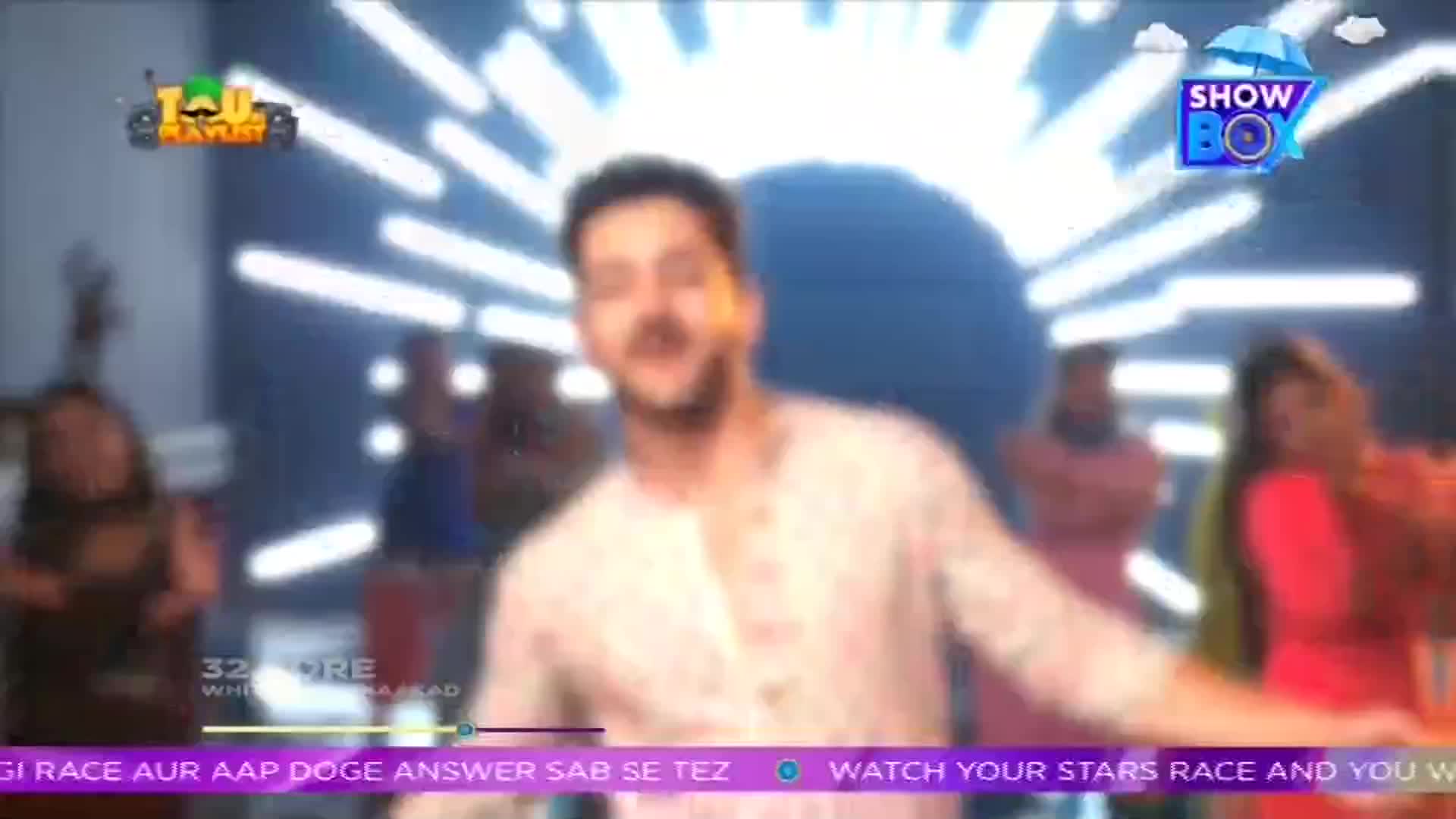 A man in a light-colored shirt gestures enthusiastically, surrounded by a group of blurred figures. The ShowBox logo sits in the upper right corner, and text at the bottom hints at a game show.
