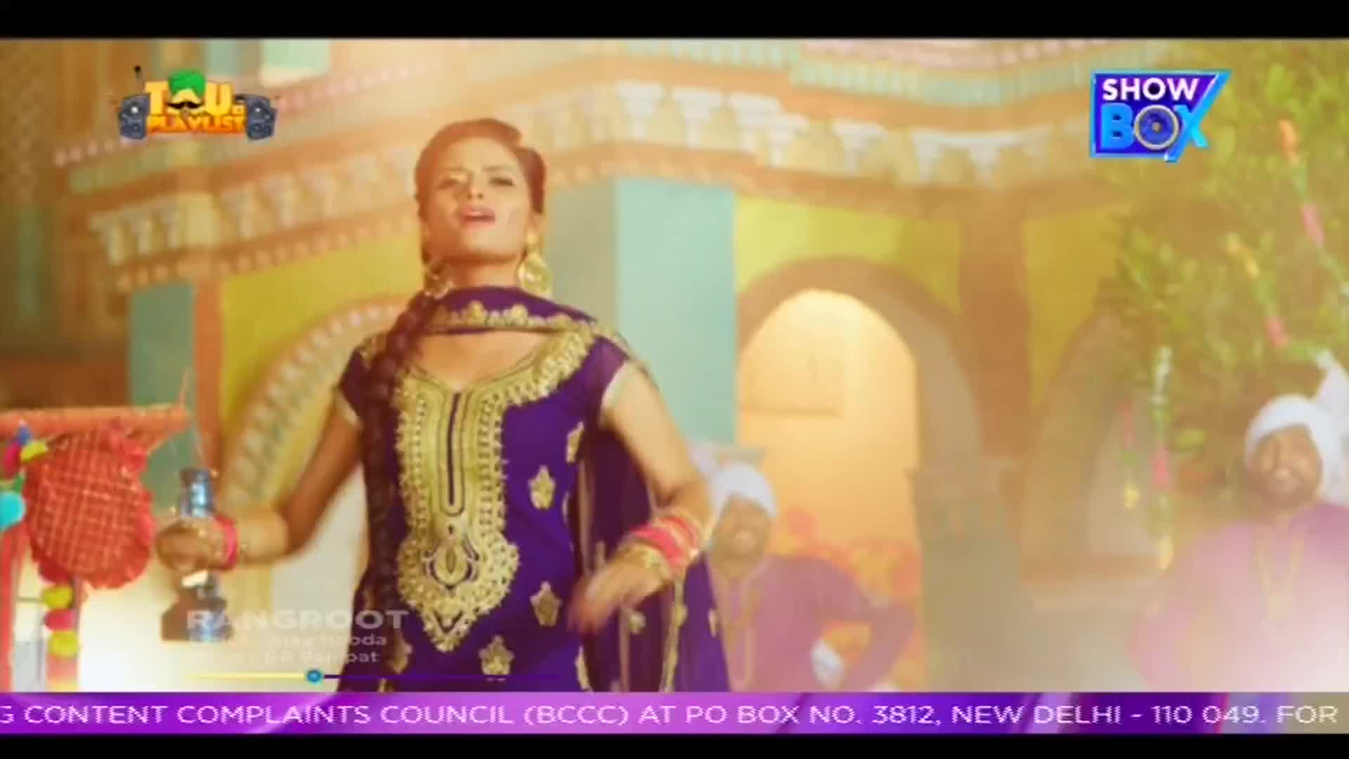 A woman in a vibrant purple and gold outfit sings, holding a small bottle. In the background, men in turbans and colorful clothing dance, all under the logo of ShowBox.

