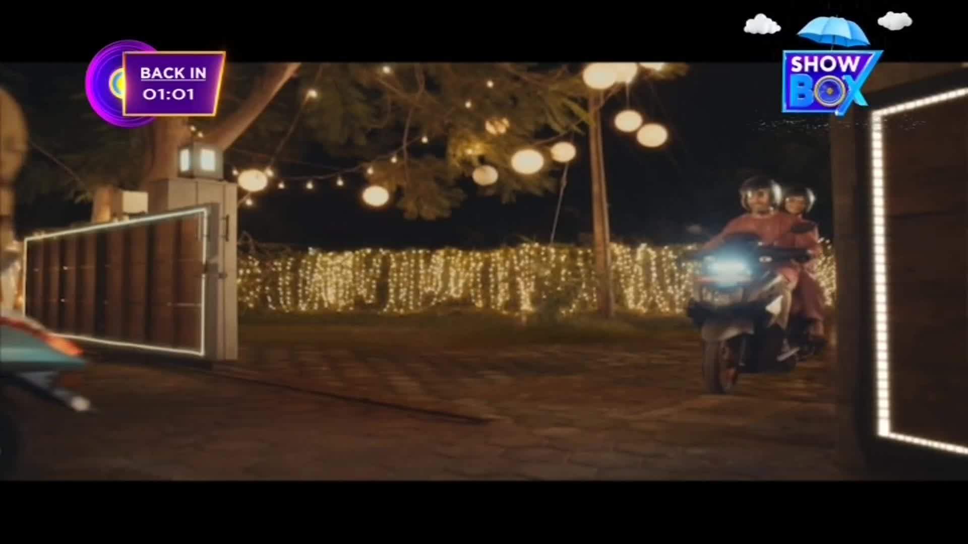 A scooter, carrying two people in helmets, is pulling up to a gate. The scene is lit by strings of lights and a ShowBox logo is visible in the corner.
