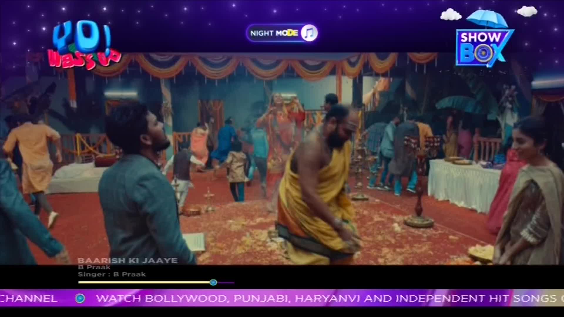 A man in a yellow robe kneels, while others in the background move around a decorated space. The scene, from a ShowBox video, feels like a celebration in India.
