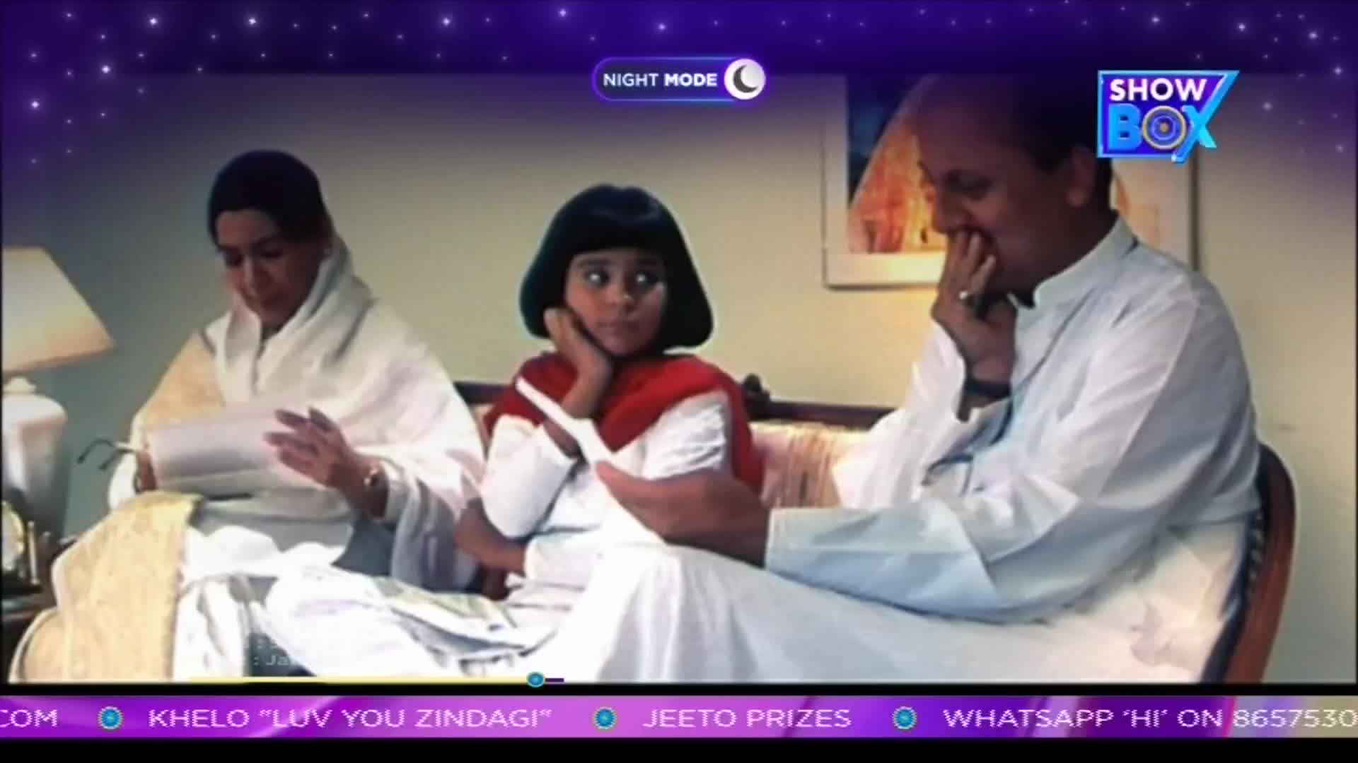 A young girl in a red scarf and white dress sits between two adults on a couch, looking off to the side. The woman on the left reads a paper, while the man on the right touches his face, as if in thought.
