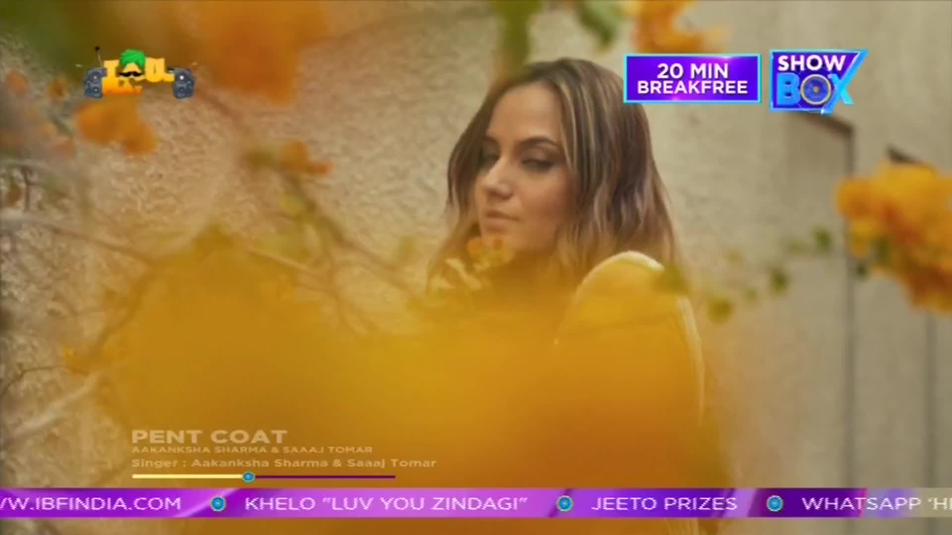 A woman with long hair is the focus, framed by yellow flowers, likely a scene from a ShowBox music video. The screen displays the song title "Pent Coat" and the names of the singers, with a "20 min Breakfree" logo in the corner.
