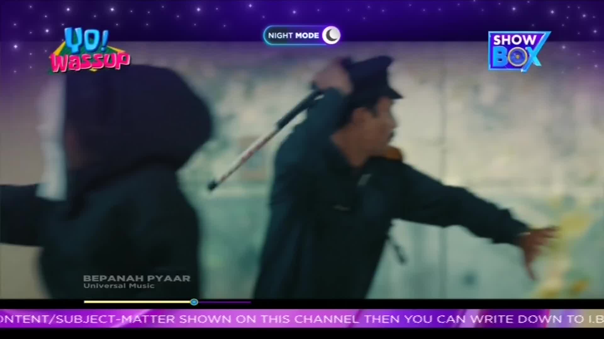 A man in a dark uniform swings a baton, his face contorted in what looks like a struggle. The scene, part of a music video on ShowBox, is set against a backdrop of what appears to be a wall.
