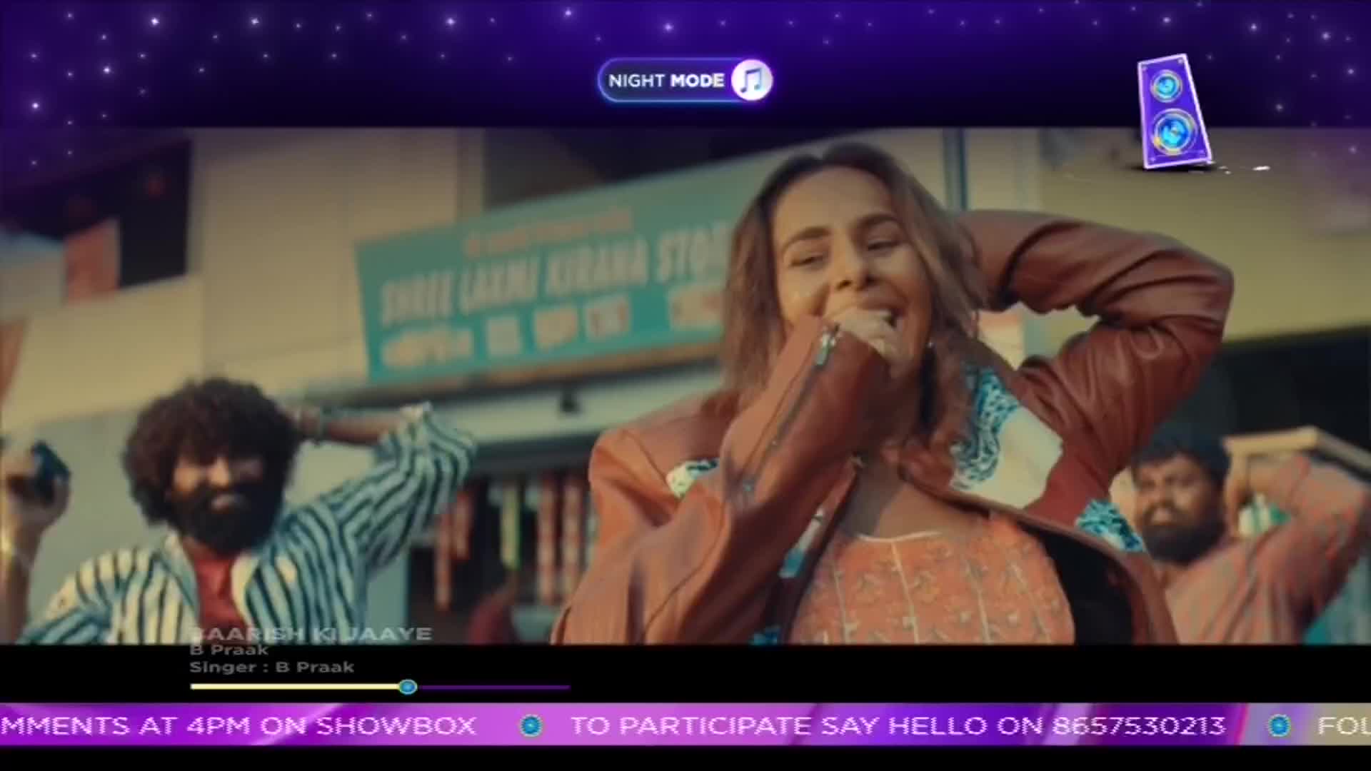 A woman in a leather jacket laughs, singing into the camera. Two men in the background, one with a striped shirt, gesture along to the music.
