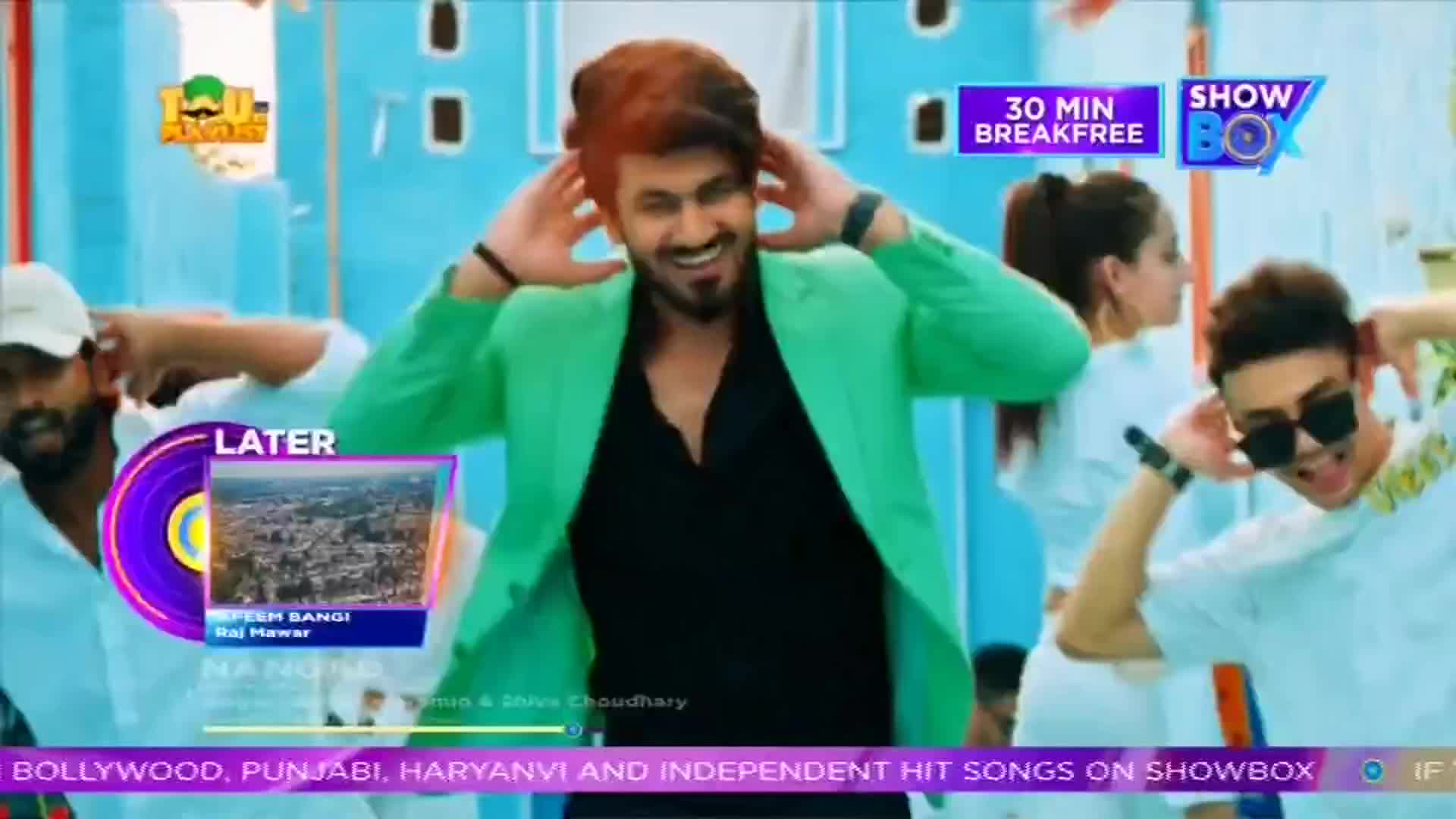 A man in a bright green jacket and a black shirt grins, his hands framing his head. Other people in white shirts and sunglasses dance behind him, all on the set of what looks like a music video on ShowBox from India.
