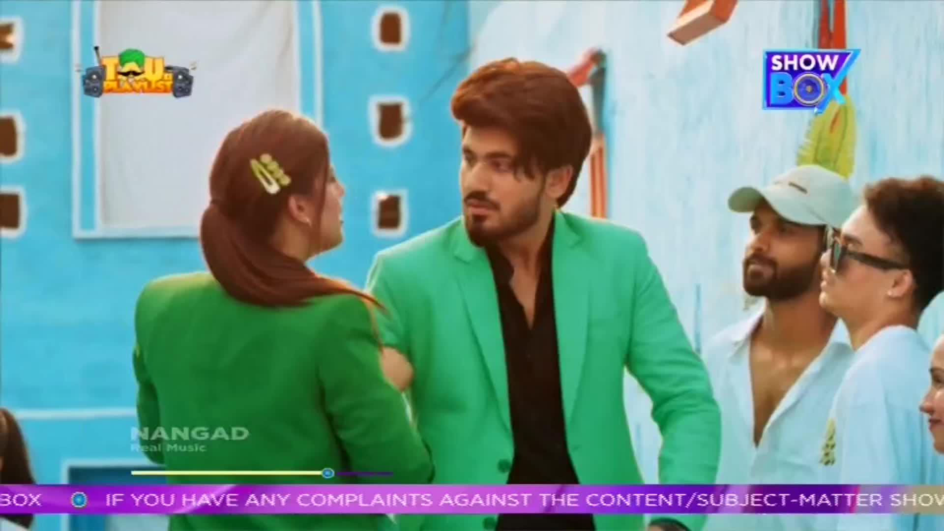 A man in a bright green suit looks intently at a woman in a matching jacket, while others stand nearby. The scene is set outdoors, with a blue wall in the background, and the logos of ShowBox and Nangad are visible.
