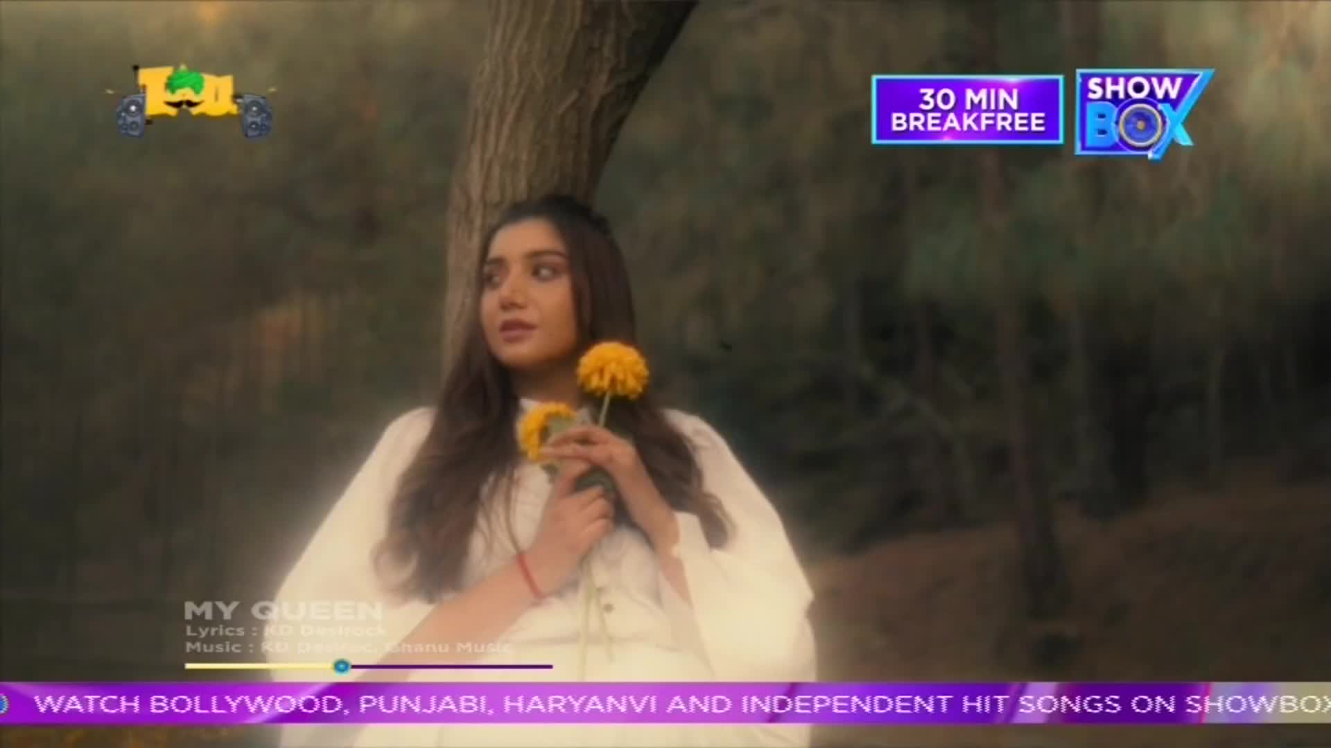 A young woman in a white dress stands holding a yellow flower, looking off to the side. The scene is framed by a tree and a ShowBox logo, with text advertising Bollywood and Punjabi music.
